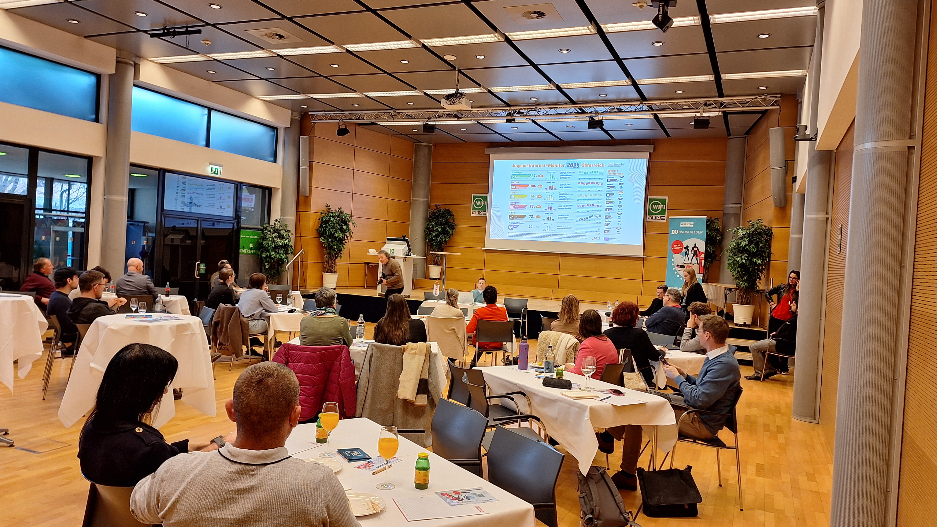 Menschen sitzen um Tische herum und lauschen einem Vortrag