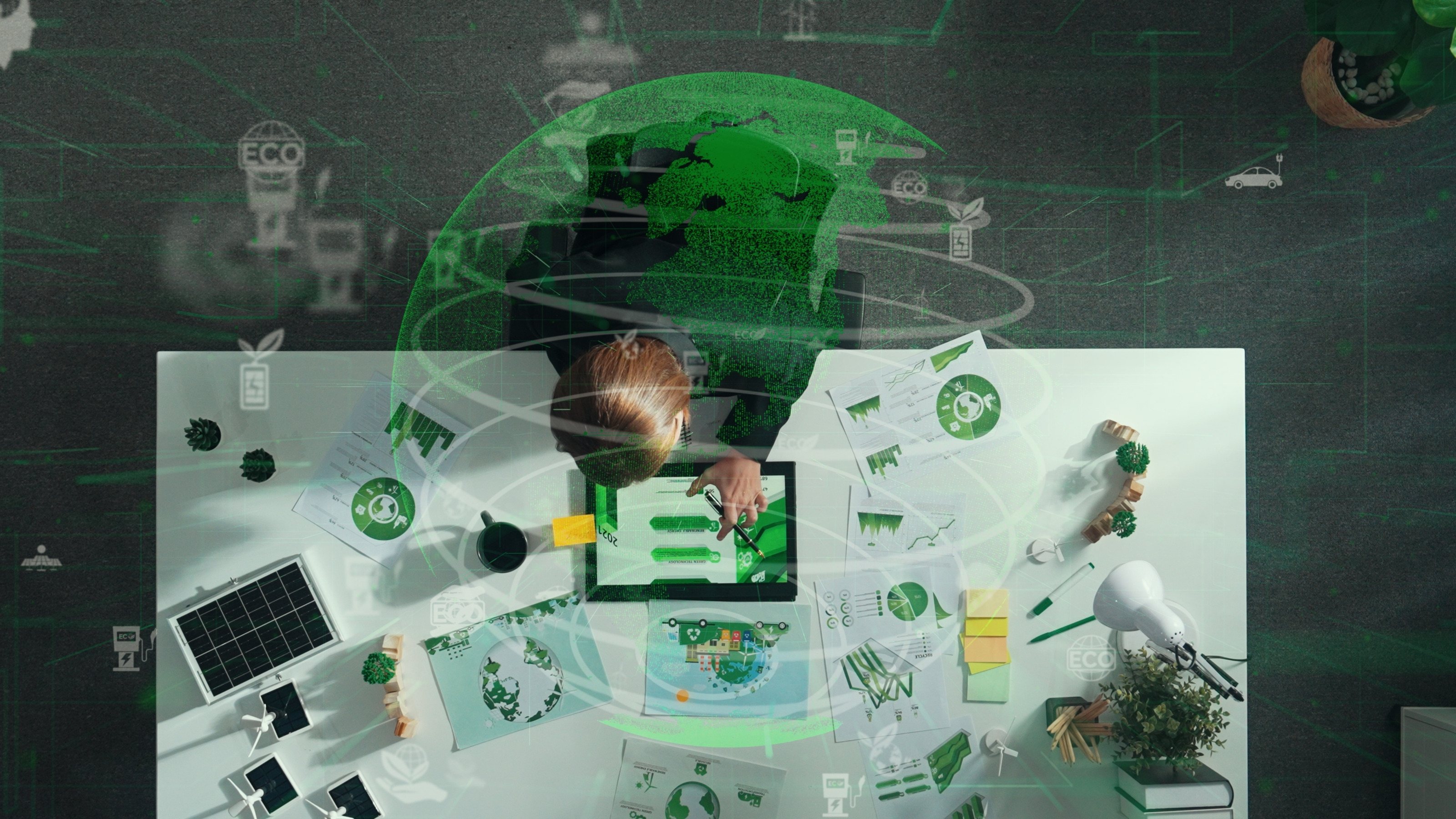 Aufnahme einer Person, die an einem Schreibtisch sitzt. Vor der Person liegt ein Tablet, auf dem gr&uuml;ne Balken sind. Daneben sind verschiedene Zettel. &Uuml;ber der Person ist eine Grafik eines Globus, in dem die Kontinente gr&uuml;n eingezeichnet sind