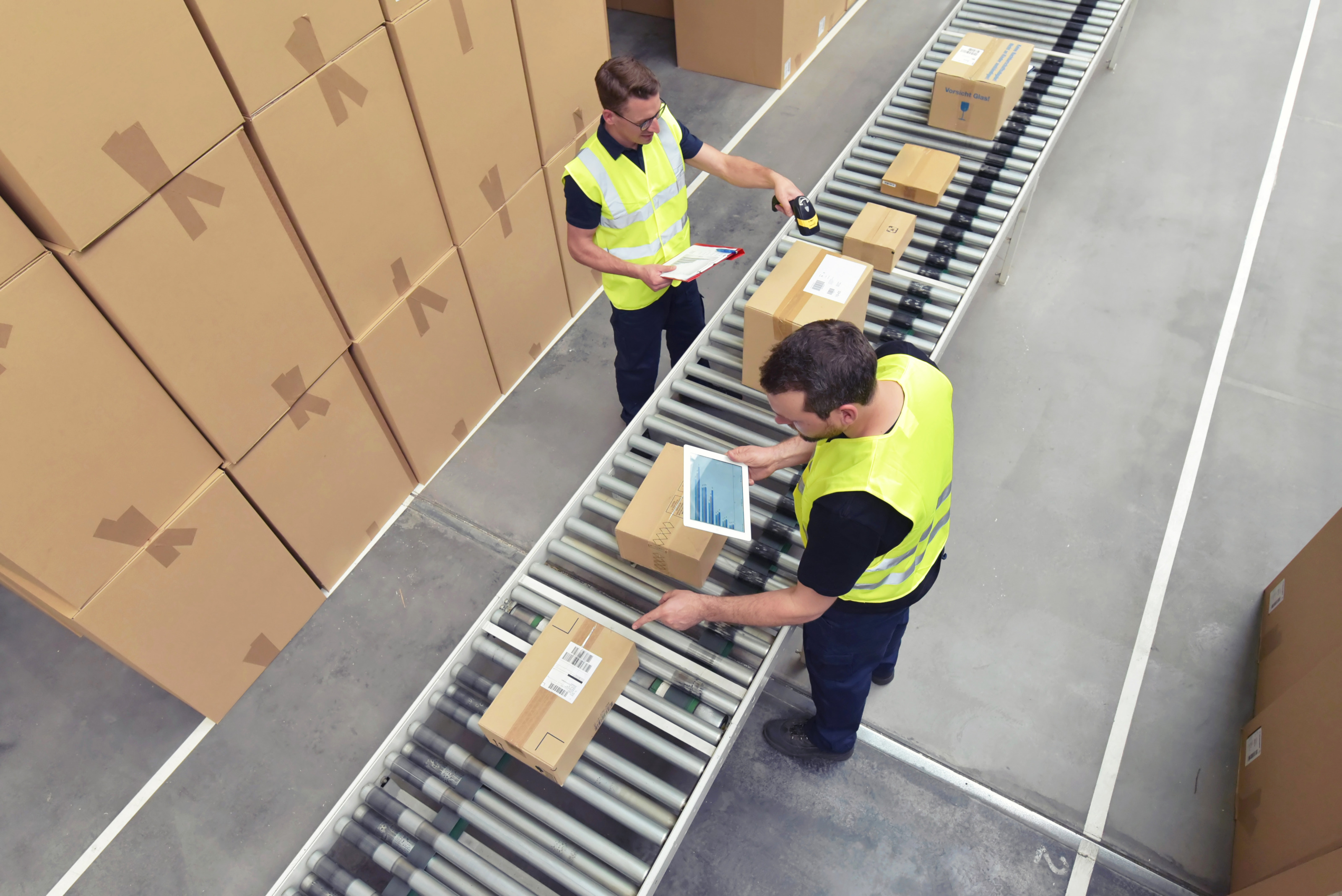 Aufnahme in Vogelperspektive: Zwei Personen in der Logistik scannen Pakete auf einem Flie&szlig;band und f&uuml;hren Statistiken.
