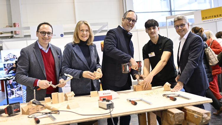 F&uuml;nf Personen an einem Hands-on-Stand der Lehrlingsmesse, darunter ein Jugendlicher und Vertreter:innen der Industrie, beim gemeinsamen Arbeiten mit Holz und Werkzeug.