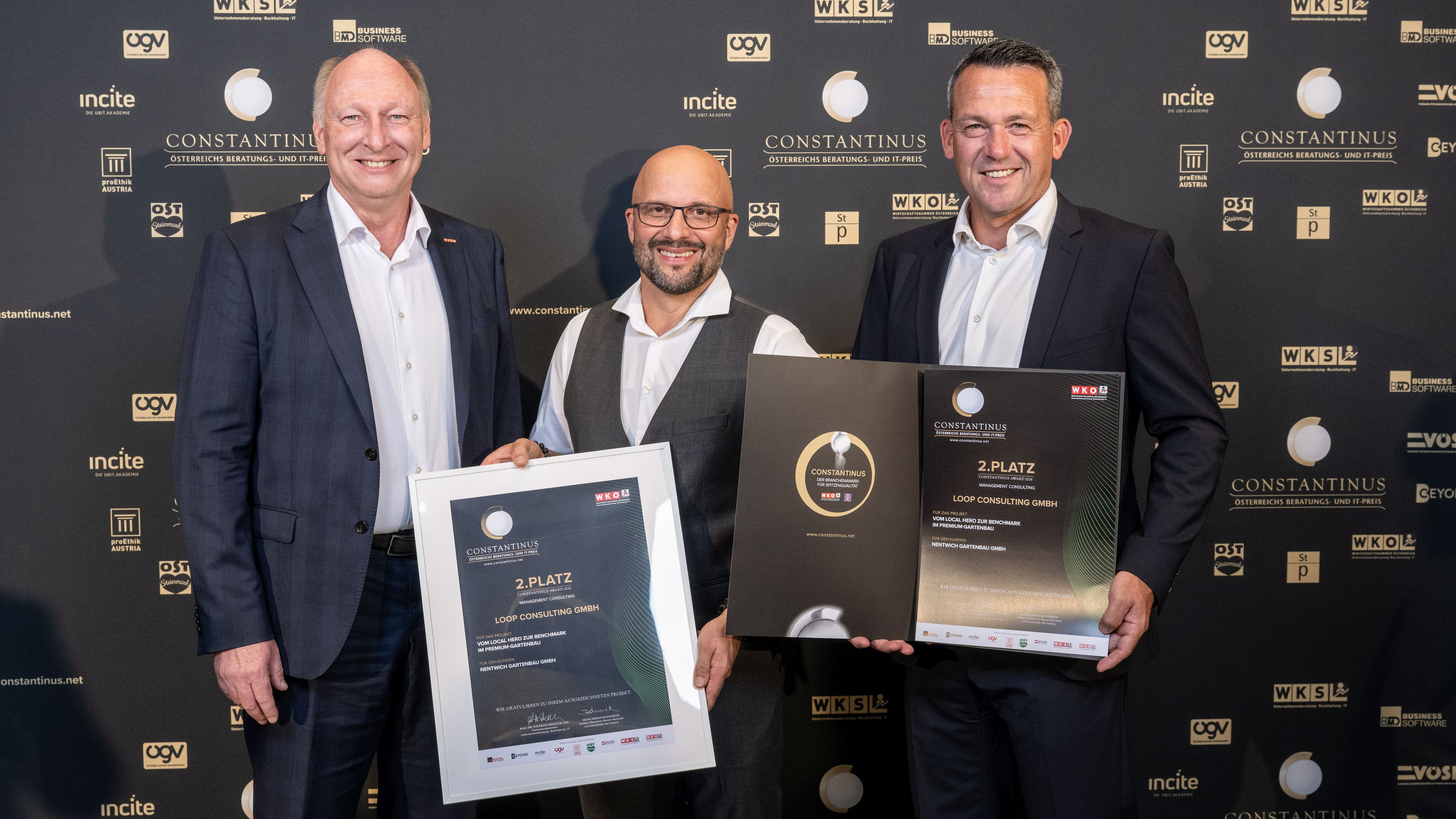 Drei M&auml;nner stehen l&auml;chelnd vor einer Constantinus-Award-Fotowand. Zwei von ihnen pr&auml;sentieren Urkunden und eine Auszeichnung f&uuml;r den 2. Platz.