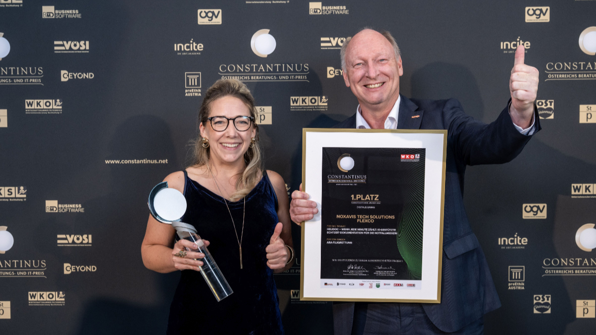 Eine Frau und ein Mann stehen l&auml;chelnd vor einer Constantinus-Award-Fotowand. Die Frau h&auml;lt eine Troph&auml;e, der Mann zeigt den Daumen nach oben und pr&auml;sentiert eine gerahmte Urkunde f&uuml;r den 1. Platz.