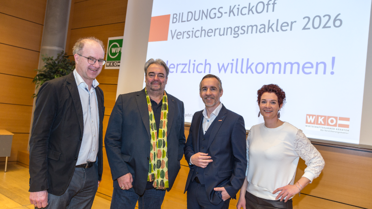 Drei M&auml;nner und eine Frau im Businessoutfit stehen auf einer B&uuml;hne - im Hintergrund sieht man eine Leinwand mit dem Schriftzug Bildungs-KickOff Versicherungsmakler, darunter steht Herzlich willkommen