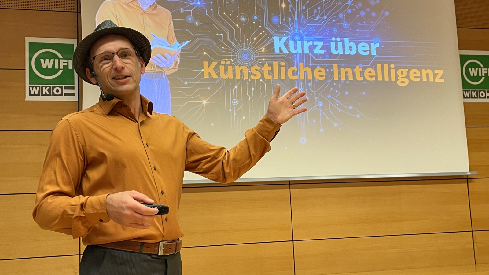 Mann mit orangem Hemd und Hut steht auf einer Bühne - im Hintergrund erkennt man den Schriftzug "Kurz über Künstliche INtelligenz"