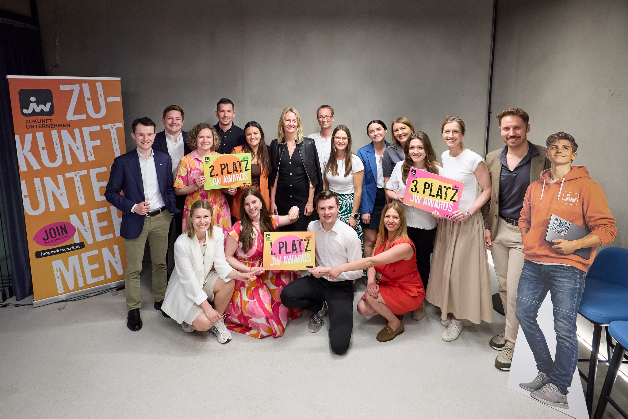Gruppenfoto von mehreren Personen vor einer grauen Wand. Drei Personen in der vorderen Reihe halten Schilder mit den Aufschriften &bdquo;1. Platz JW Awards&ldquo;, &bdquo;2. Platz JW Awards&ldquo; und &bdquo;3. Platz JW Awards&ldquo;. Links steht ein Roll-up-Banner mit dem JW-Logo