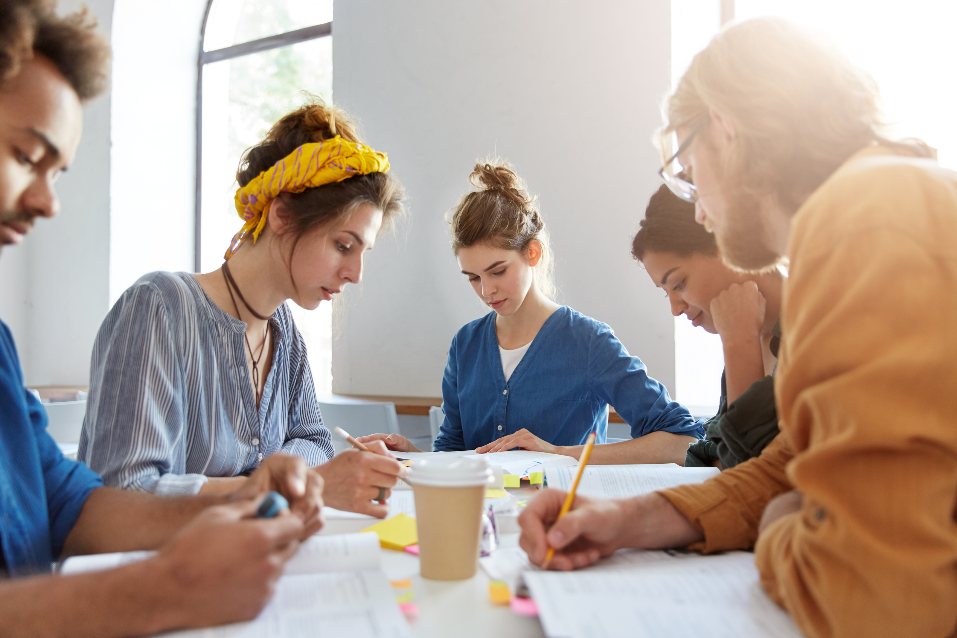 Jugendliche Personen sitzen gemeinsam bei einem Tisch, blicken auf Unterlagen und lernen