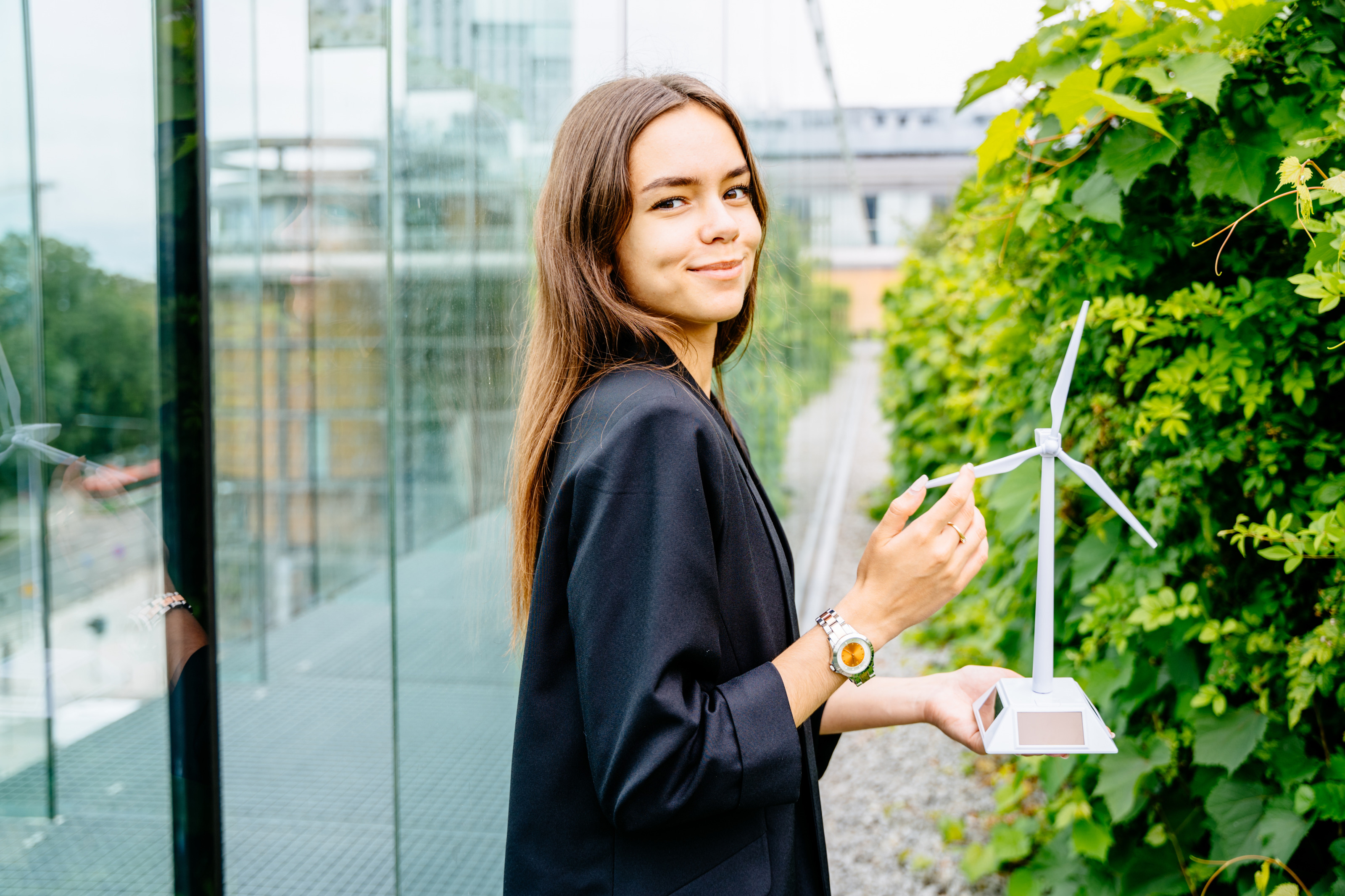 Sanft l&auml;chelnde Person h&auml;lt Modell eines Windrades in H&auml;nden, zwischen Glasfassade eines Geb&auml;udes und gr&uuml;ner Hecke stehend