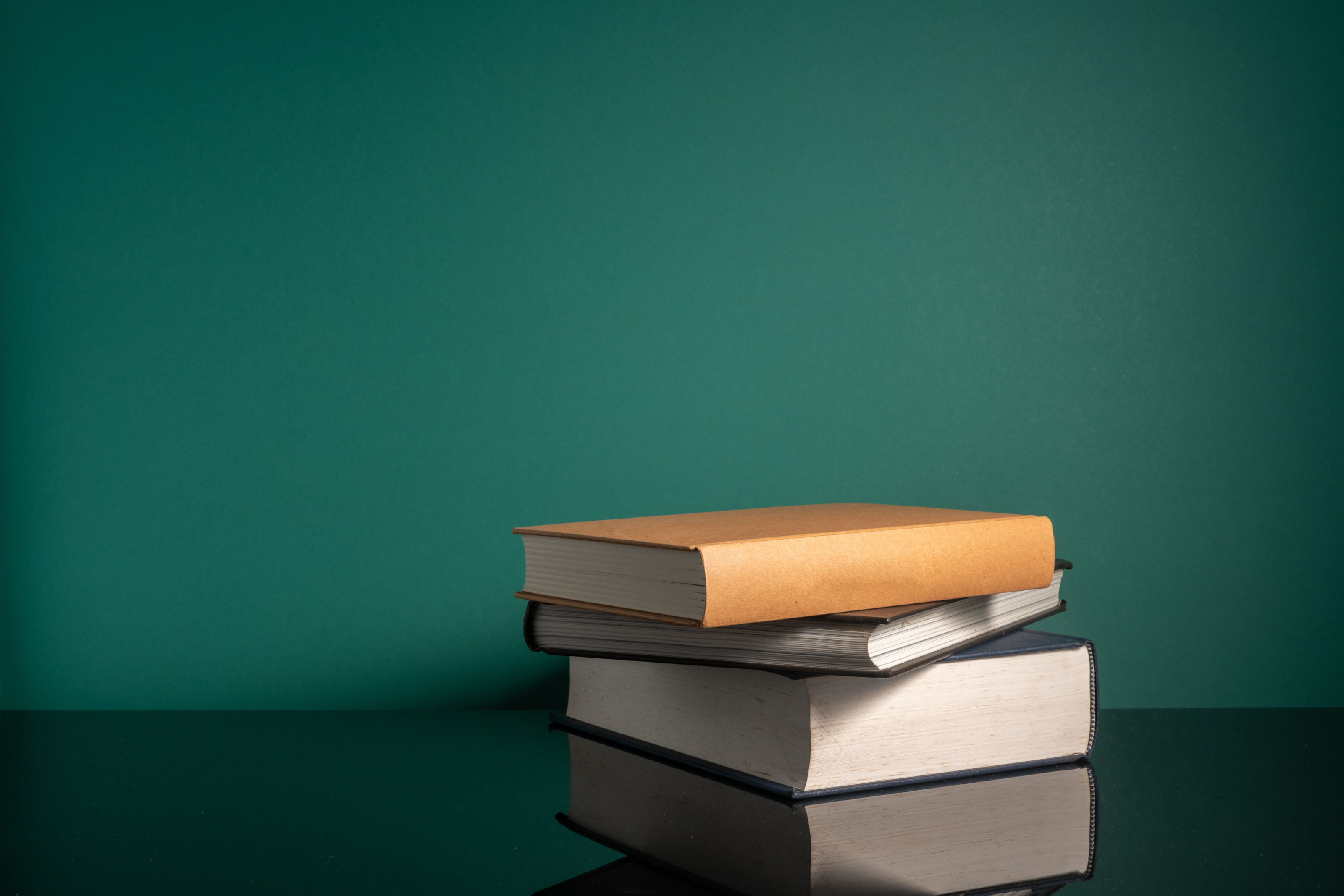 Stapel aus drei Hardcoverb&uuml;chern, der sich auf darunterliegendem Untergrund spiegelt vor gr&uuml;ner Wand