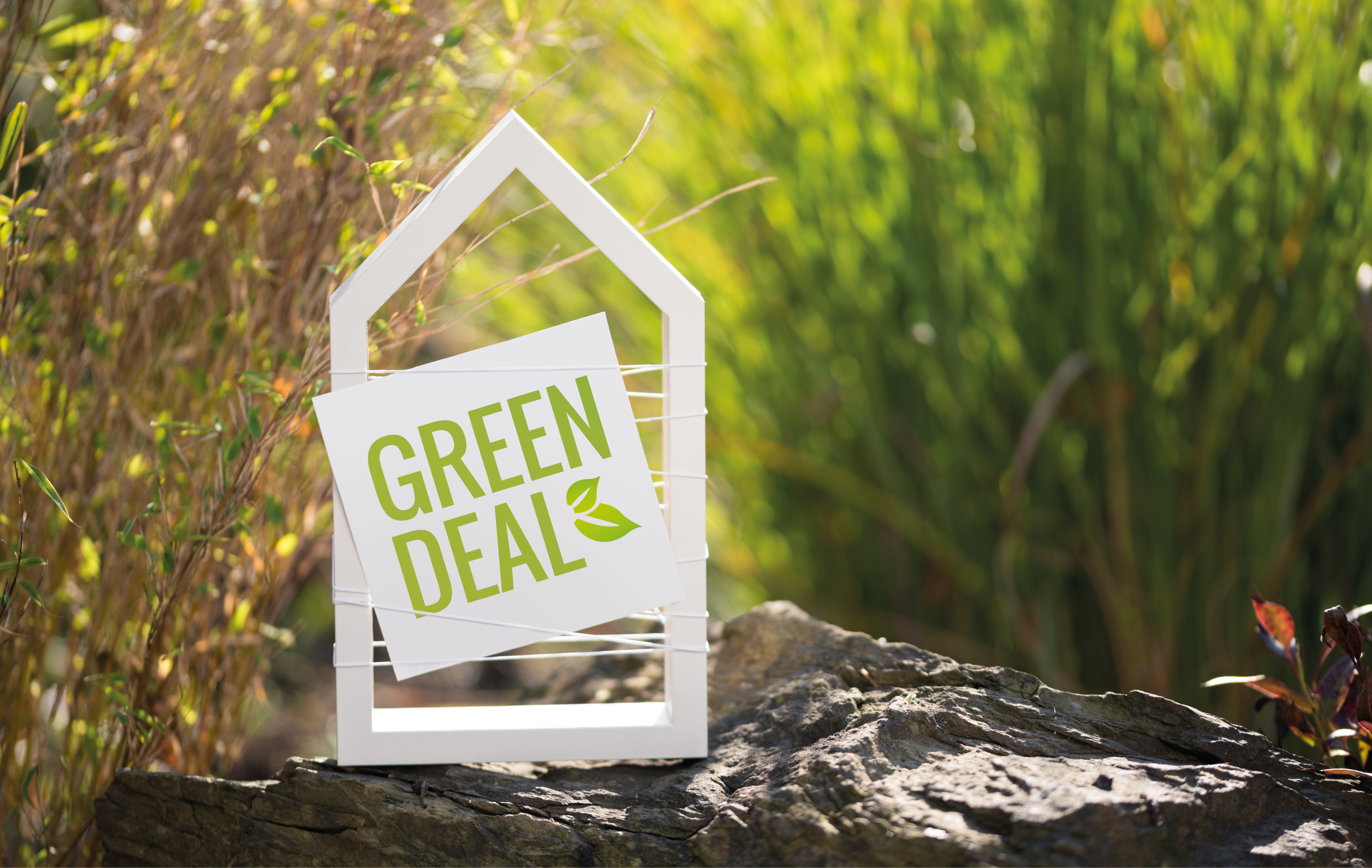 Wei&szlig;e Form eines H&auml;uschen mit Schild mit gr&uuml;ner Schrift und Symbol eines Blattes Green Deal auf Stein platziert, im Hintergrund verschwommen gr&uuml;nbraun Schilf