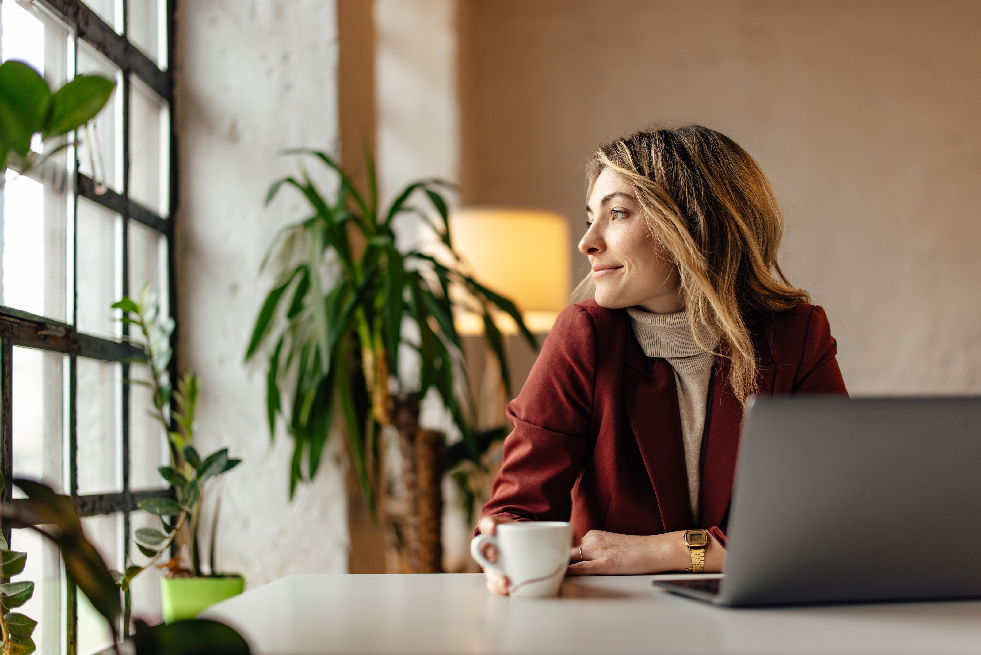 Sanft l&auml;chelnde Person an Schreibtisch vor aufgeklapptem Laptop sitzend, h&auml;lt wei&szlig;e Tasse in einer Hand und blickt zur Seite aus Fenster, im Hintergrund verschwommen Zimmerpflanzen und Stehleuchte