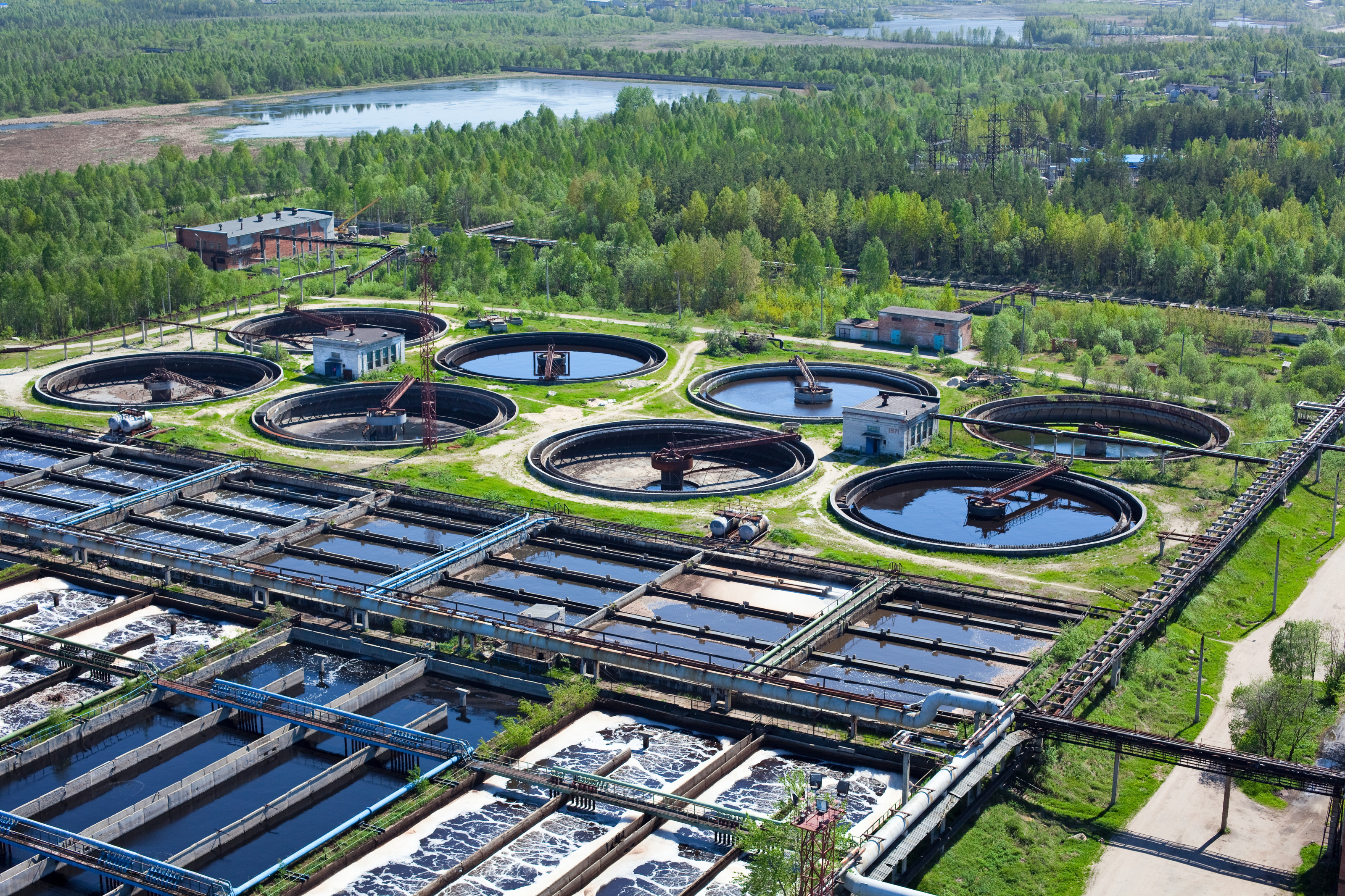 Vogelperspektive einer Wasseraufbereitungsanlage: Runde und viereckige Wasserbecken nebeneinander in gr&uuml;ner Landschaft mit B&auml;umen