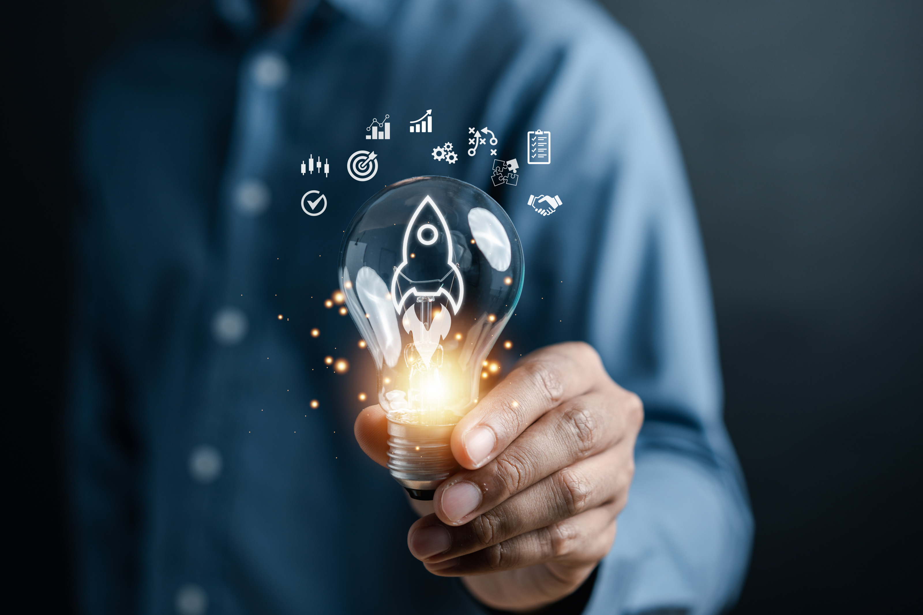 Eine Person hält eine Glühbirne in die Kamera. In der Glühbirne sind die Umrisse einer Rakete. Am unteren Ende ist die Glühbirne beleuchtet. Über der Glühbirne sind grafische Symbole wie eine Zielscheibe, Zahnräder, eine Checkliste oder Puzzleteile