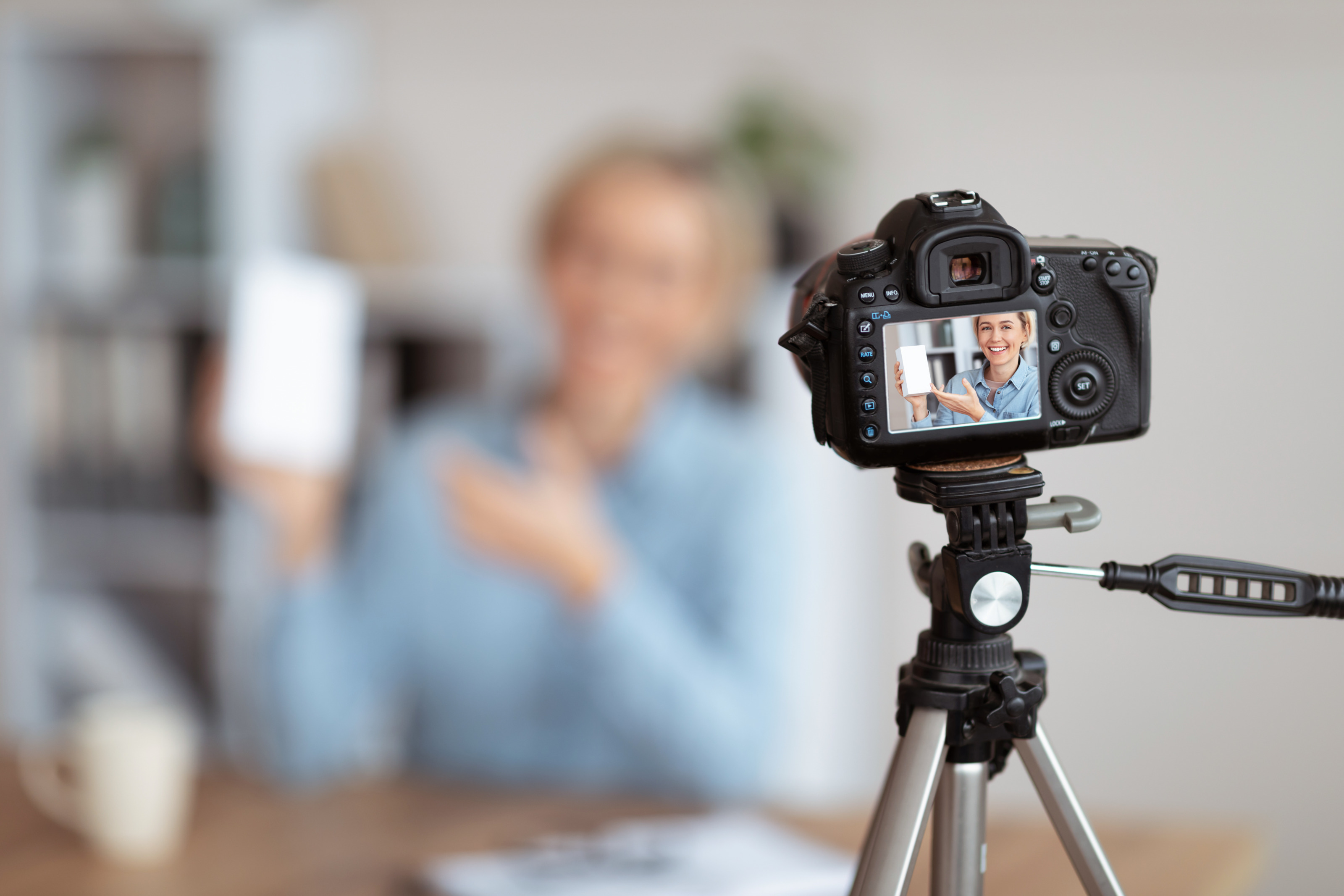 Im Vordergrund ist die Rückenansicht einer Kamera, die auf einem Stativ steht. Auf dem Bildschirm der Kamera ist eine Person, die eine Verpackung in die Kamera hält. Im Hintergrund sitzt diese Person an einem Tisch