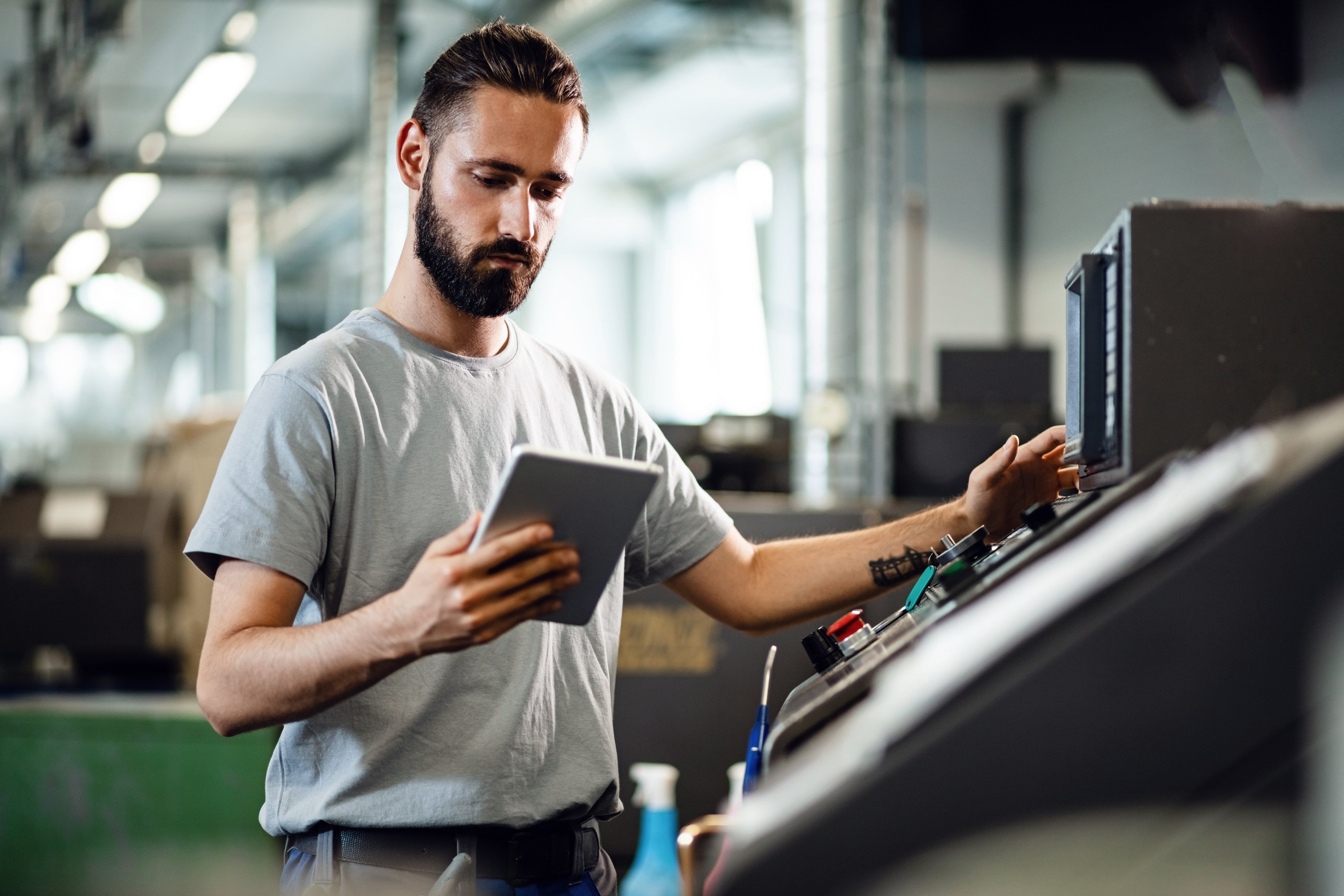 Eine Person steht vor einer CNC-Maschine und h&auml;lt ein Tablet in der rechten Hand. Die Person schaut konzentriert auf das Tablet. Im Hintergrund ist eine Industriehalle zu sehen.