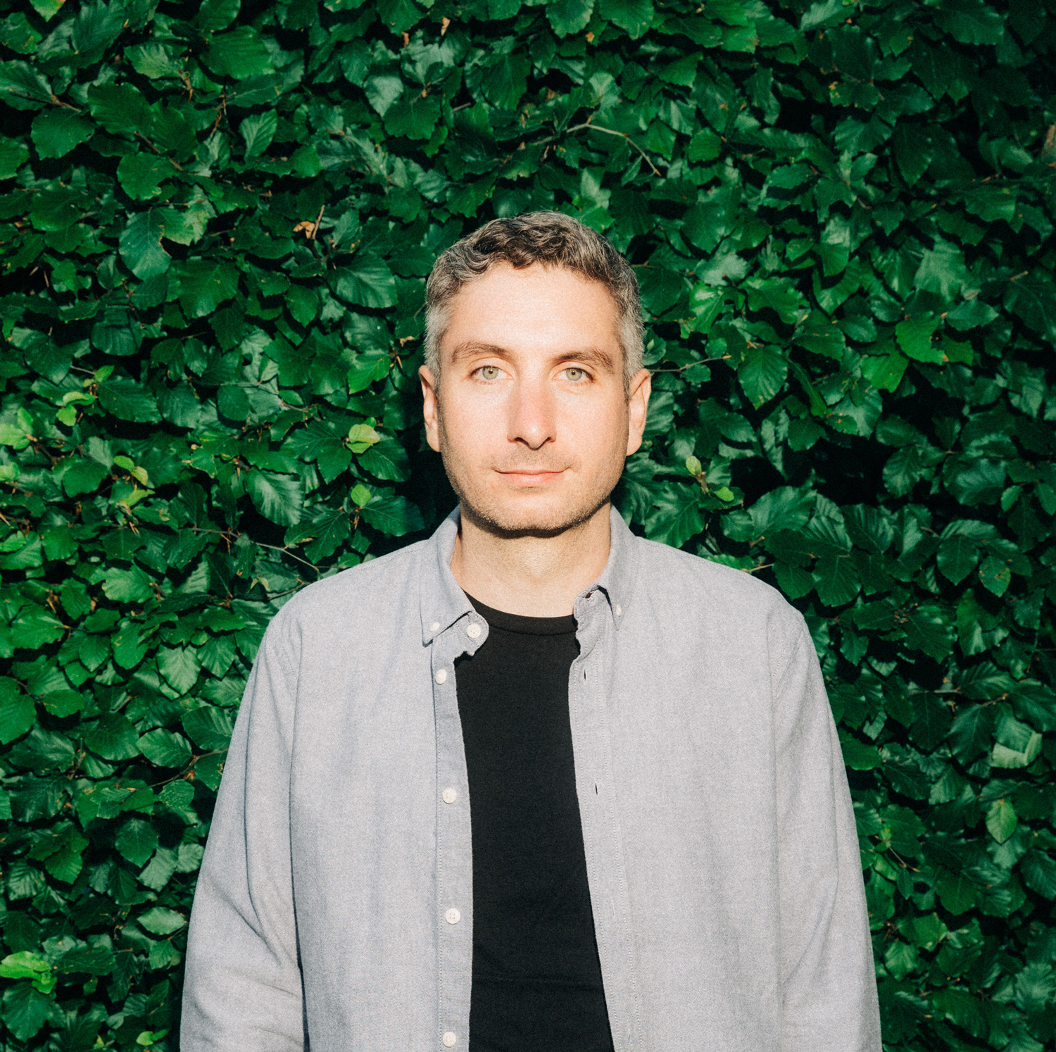 Portr&auml;t einer Person vor gr&uuml;ner Hecke, mit grauem Hemd &uuml;ber schwarzem T-Shirt