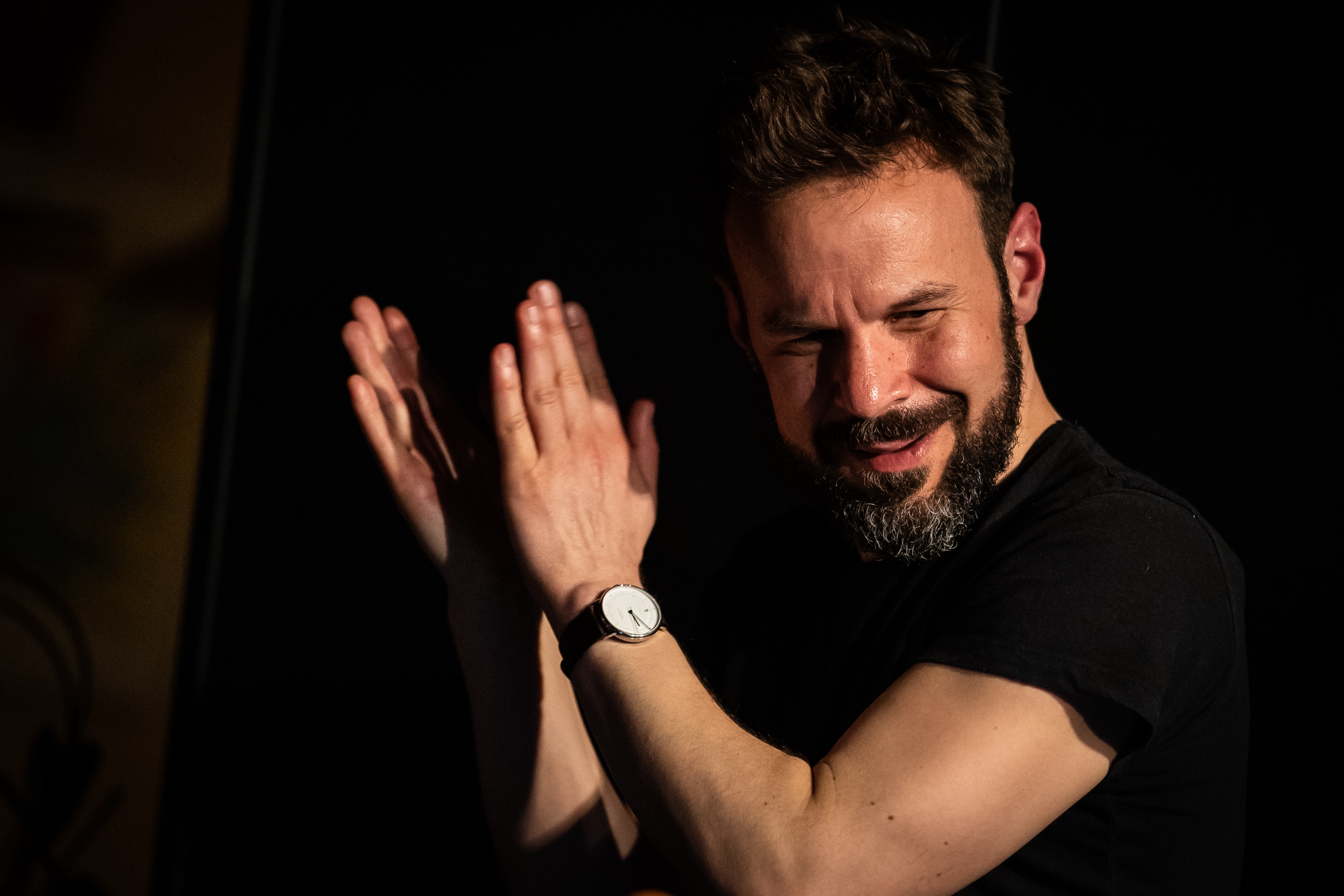 Person mit Bart klatscht in die H&auml;nde, tr&auml;gt schwarzes T-Shirt und Armbanduhr vor dunklem Hintergrund