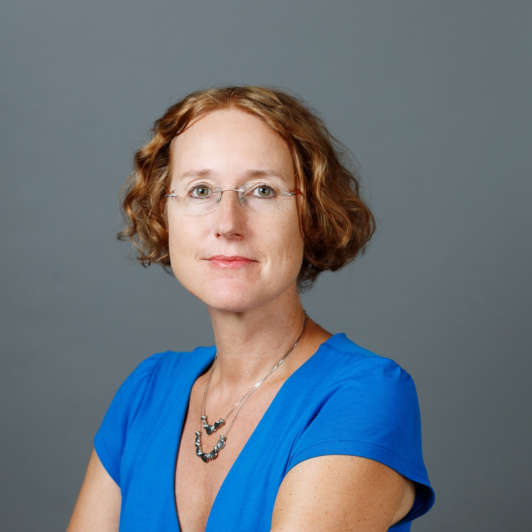 Portr&auml;tfoto: Person mit kurzen lockigen Haaren, Brille und blauem Oberteil vor grauem Hintergrund