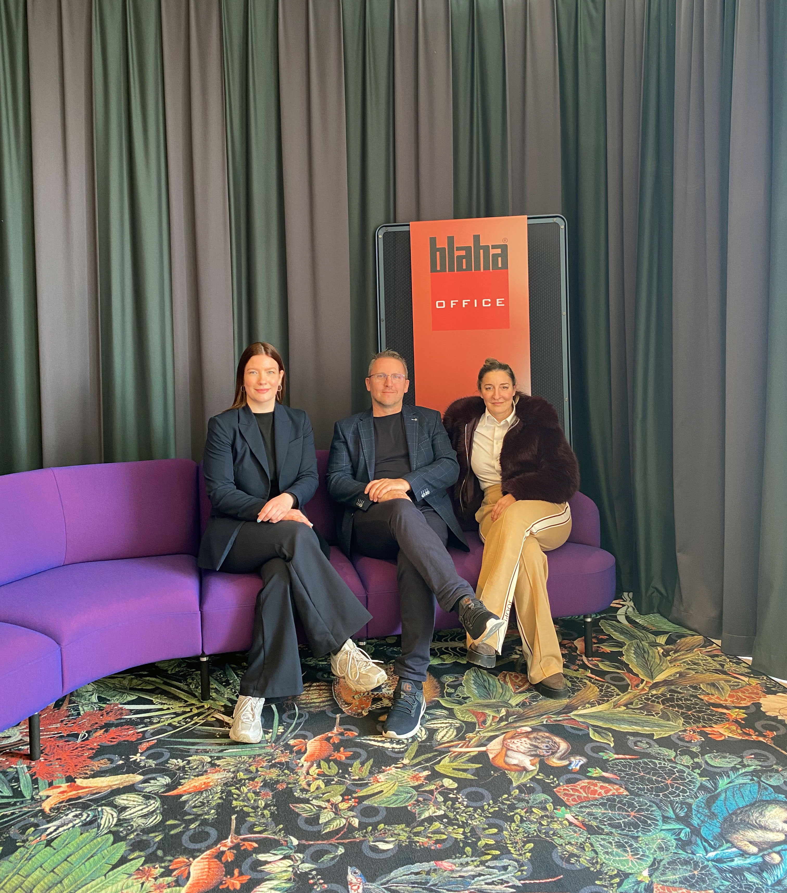 Drei Personen sitzen auf einem violetten Sofa vor einem Vorhang; im Hintergrund steht ein Schild mit der Aufschrift &bdquo;blaha OFFICE&ldquo;, am Boden liegt ein gemusterter Teppich.