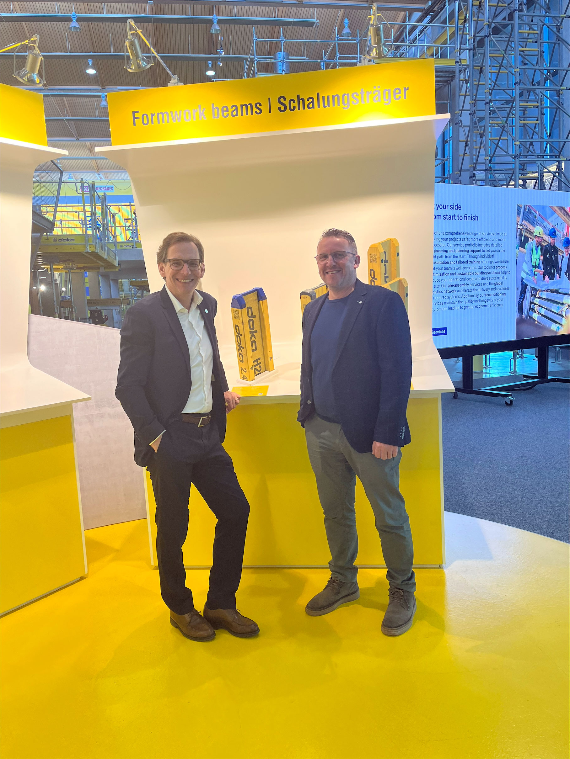 Zwei Personen stehen an einem gelben Messestand mit der Aufschrift &bdquo;Formwork beams | Schalungstr&auml;ger&ldquo;, im Hintergrund eine Messehalle mit Ger&uuml;sten und Monitor.