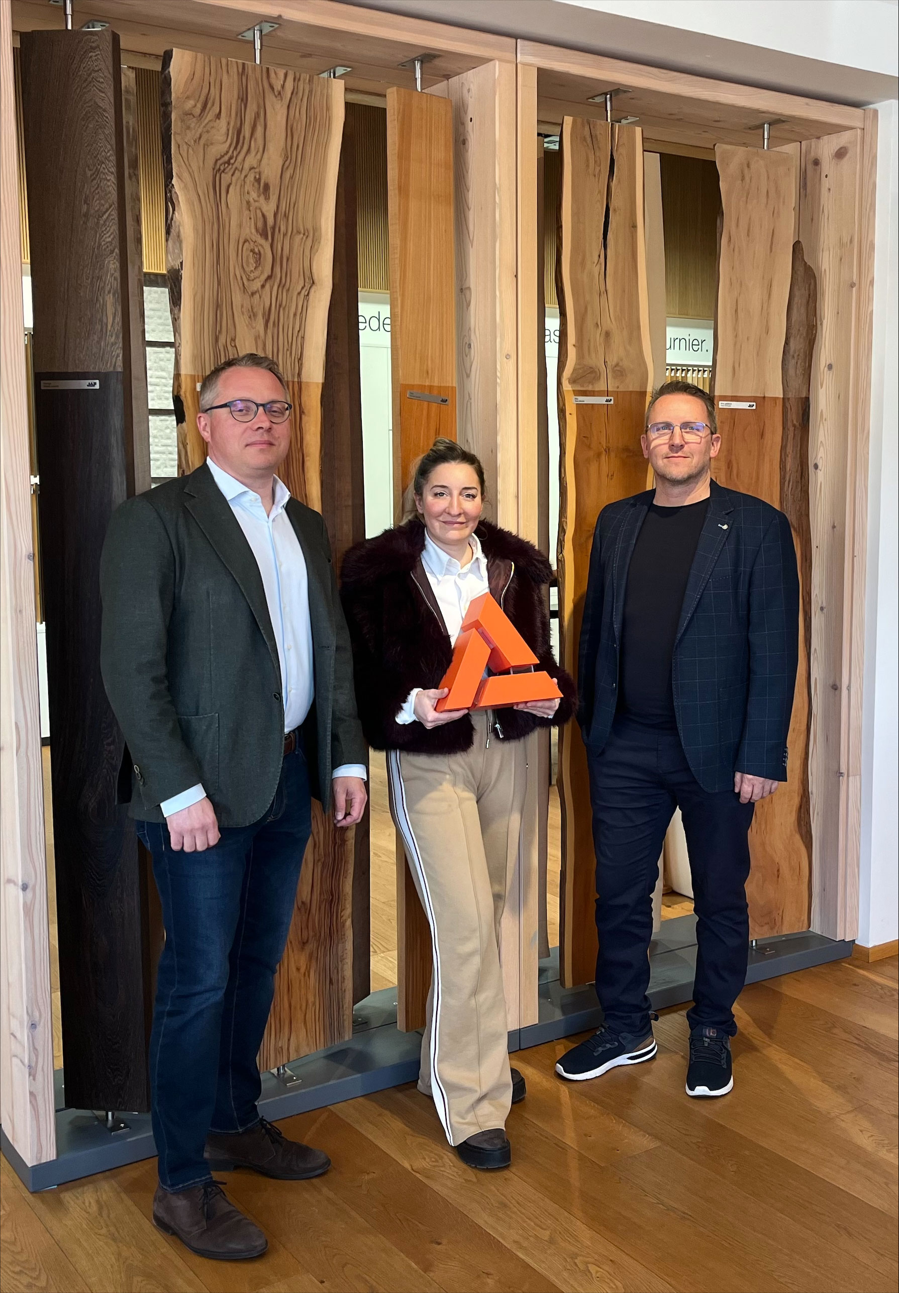 Drei Personen stehen in einem Innenraum vor einer Pr&auml;sentationswand mit verschiedenen Holzoberfl&auml;chen; eine Person h&auml;lt eine dreieckige, orangefarbene Auszeichnung in den H&auml;nden.