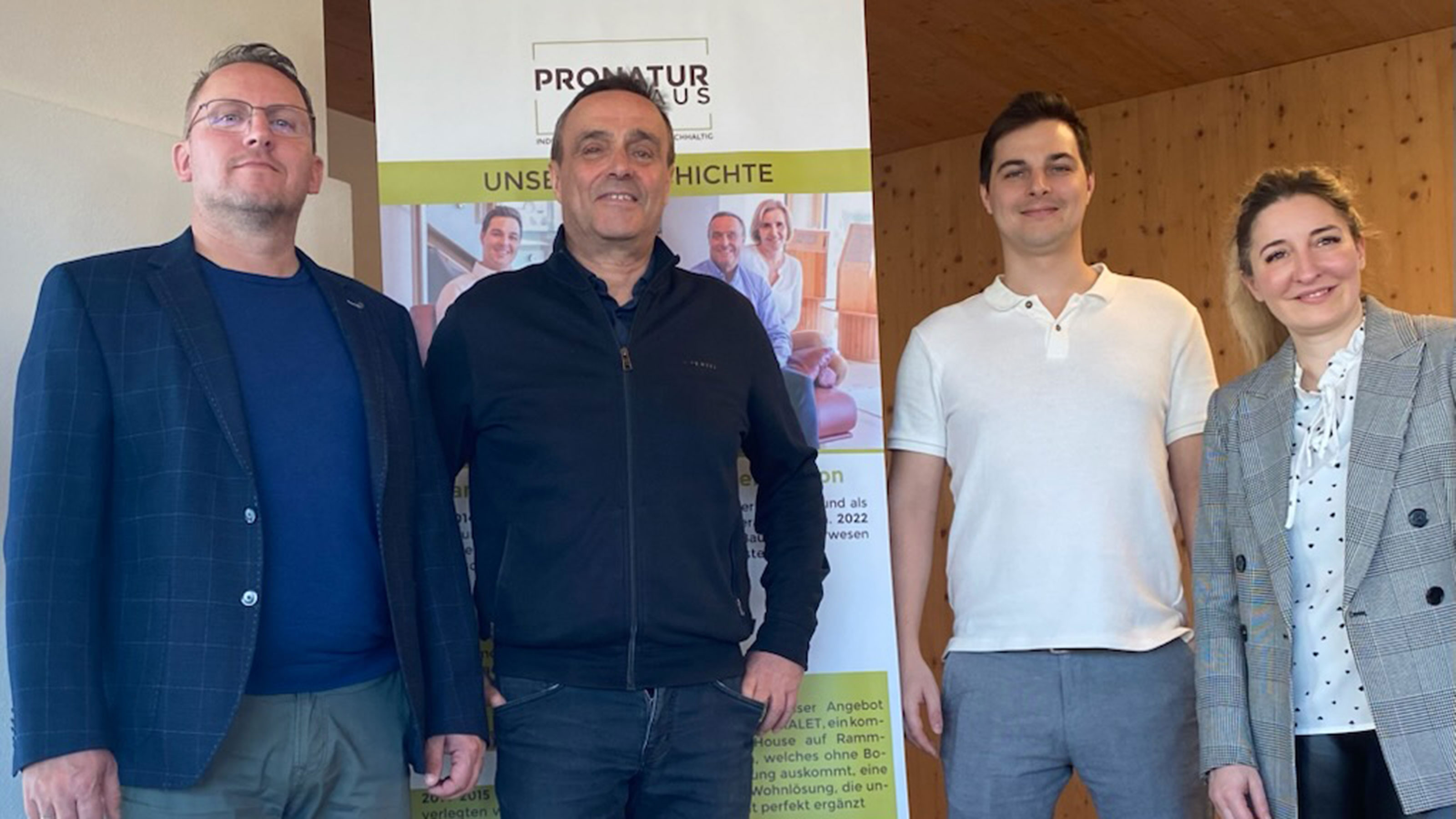 Vier Personen stehen nebeneinander in einem Innenraum vor einem Roll-up-Banner mit der Aufschrift &bdquo;PRONATUR HAUS &ndash; Unsere Geschichte&ldquo;.