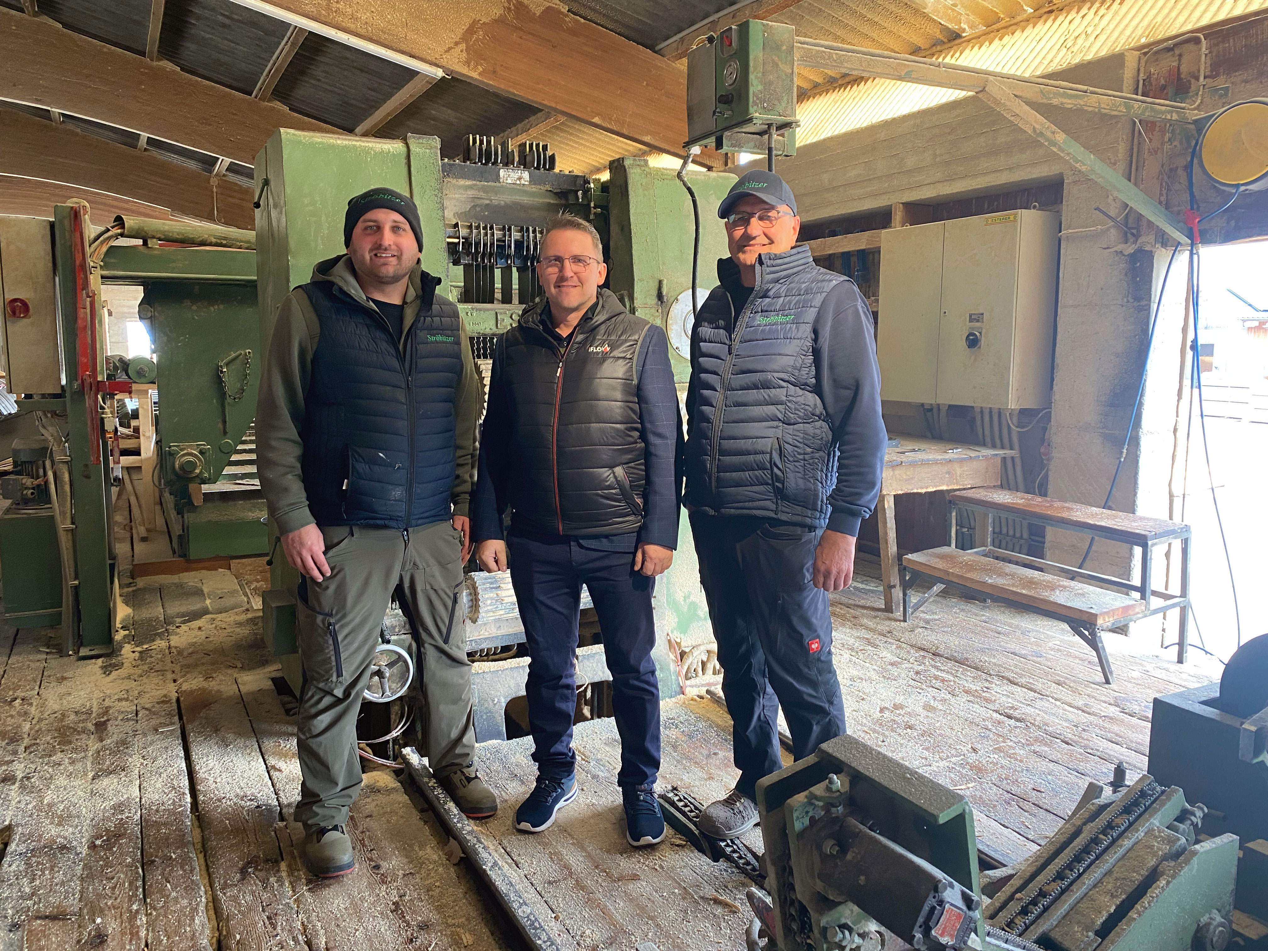 Drei Personen stehen in einer Werkhalle vor einer gro&szlig;en gr&uuml;nen Industriemaschine auf einem mit S&auml;gesp&auml;nen bedeckten Holzboden.