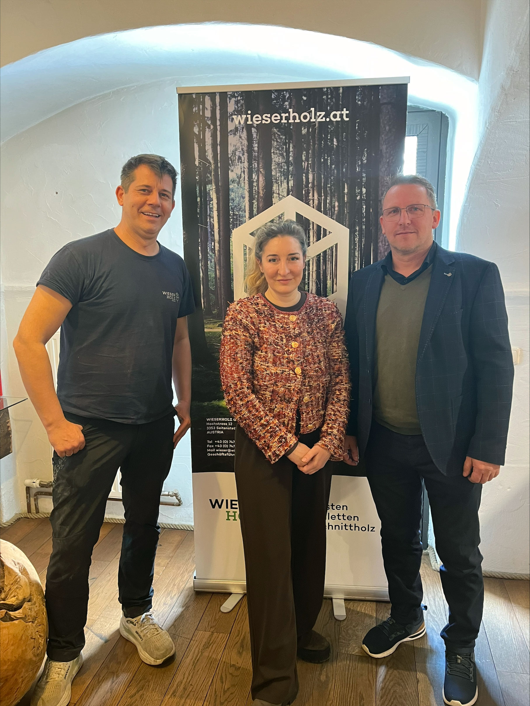 Drei Personen stehen in einem Innenraum vor einem Roll-up-Banner mit Waldmotiv und der Aufschrift &bdquo;wieserholz.at&ldquo;.