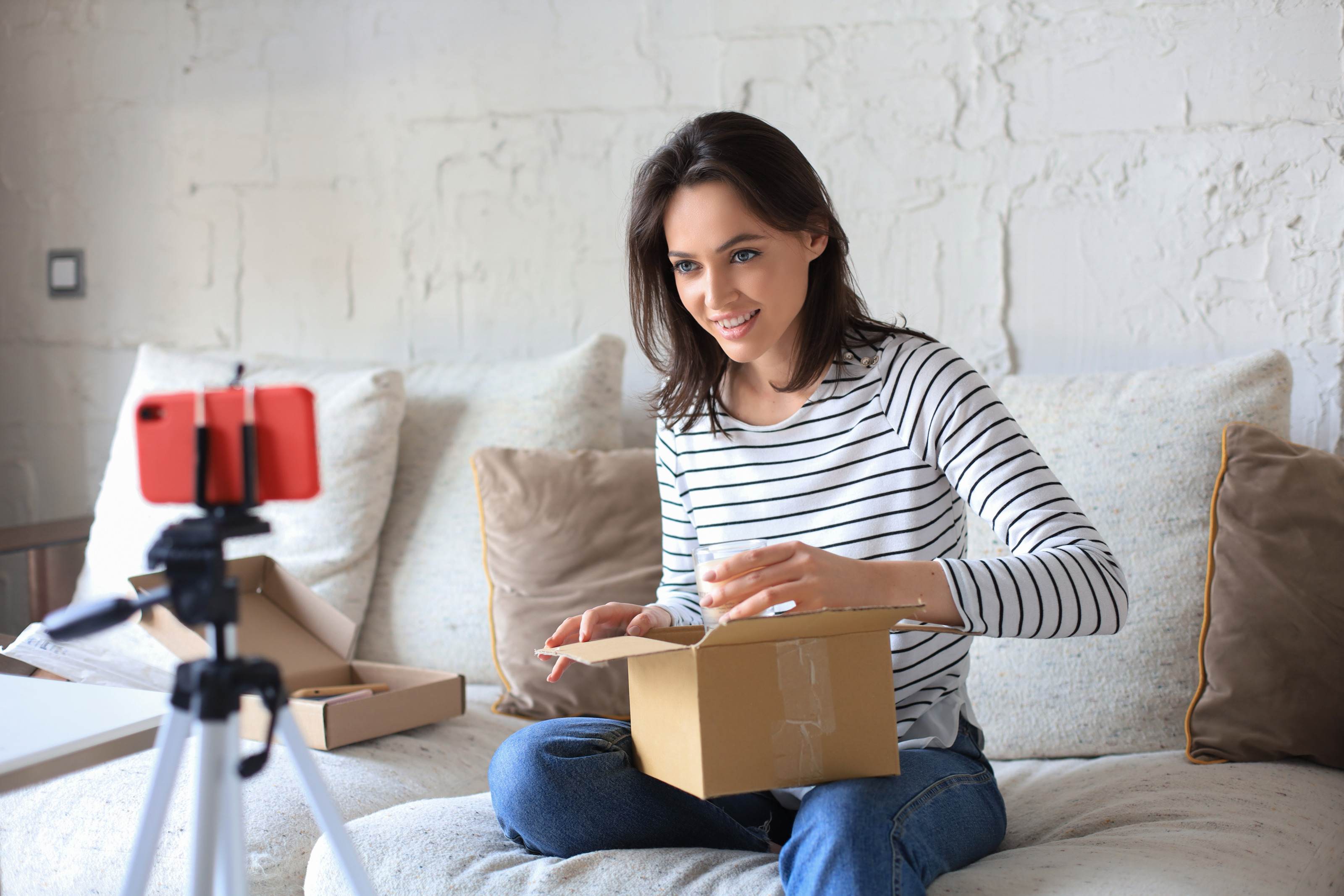 Eine Person sitzt auf einem hellen Sofa. Auf dem Scho&szlig; hat sie einen kleinen Karton, den sie &ouml;ffnet. Ihr gegen&uuml;ber ist ein Handy auf einem Stativ fixiert, das die Person beim &Ouml;ffnen filmt