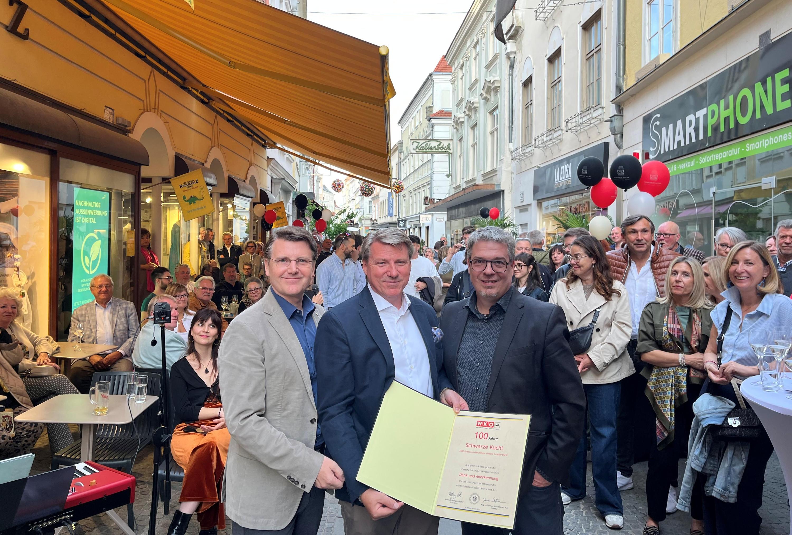 Feier in einer Fu&szlig;g&auml;ngerzone: Drei Personen pr&auml;sentieren eine WKN&Ouml;‑Urkunde &bdquo;100 Jahre Schwarze Kuchl&ldquo;, umgeben von zahlreichen Besucher:innen und Luftballons vor Gesch&auml;ften und Gastgarten.