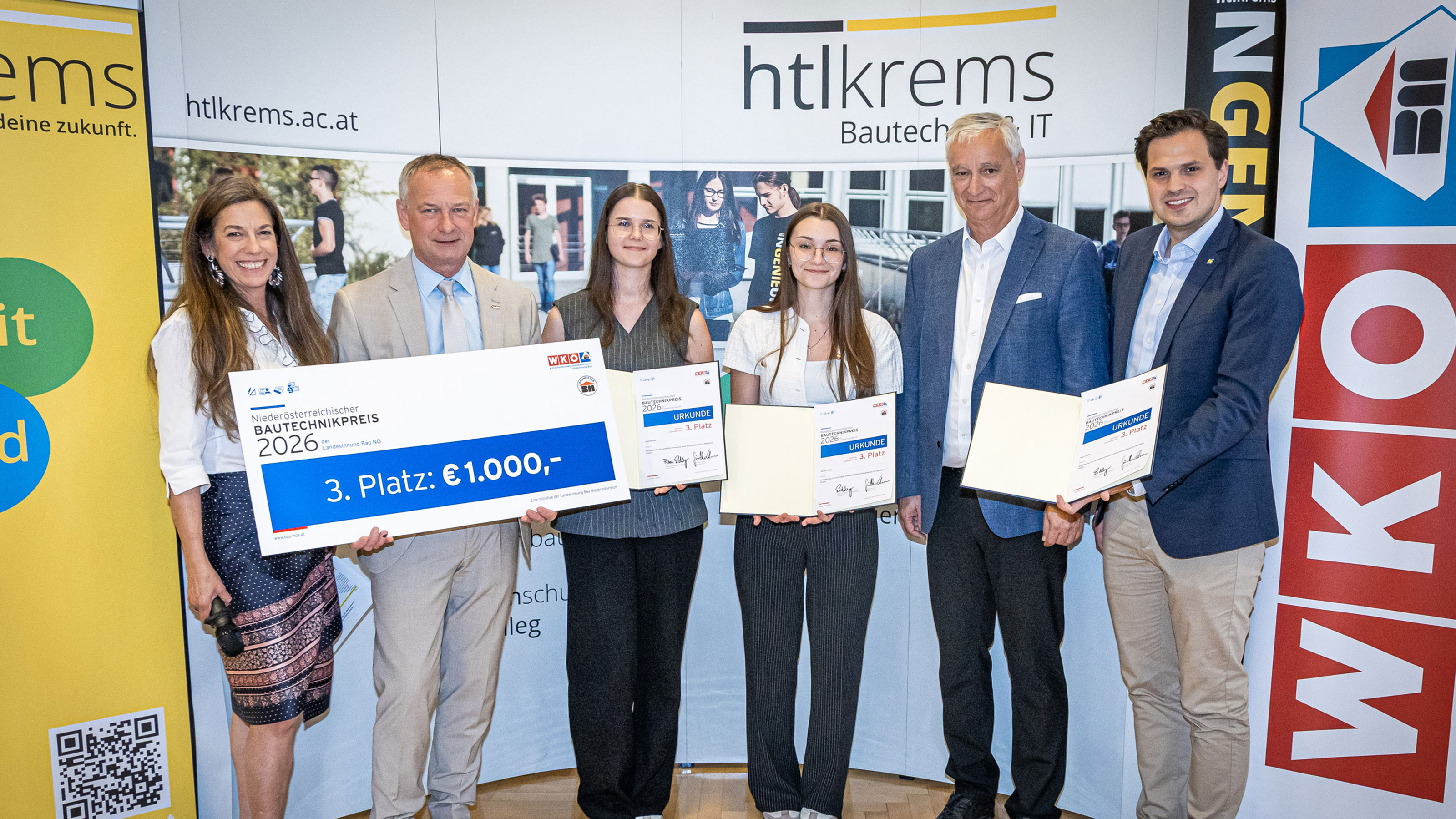 Gruppe von Personen bei einer Preisverleihung vor einer HTL‑Krems‑Fotowand, eine Person h&auml;lt einen Scheck mit der Aufschrift Bautechnikpreis 2026 &ndash; 3. Platz: 1.000 Euro.