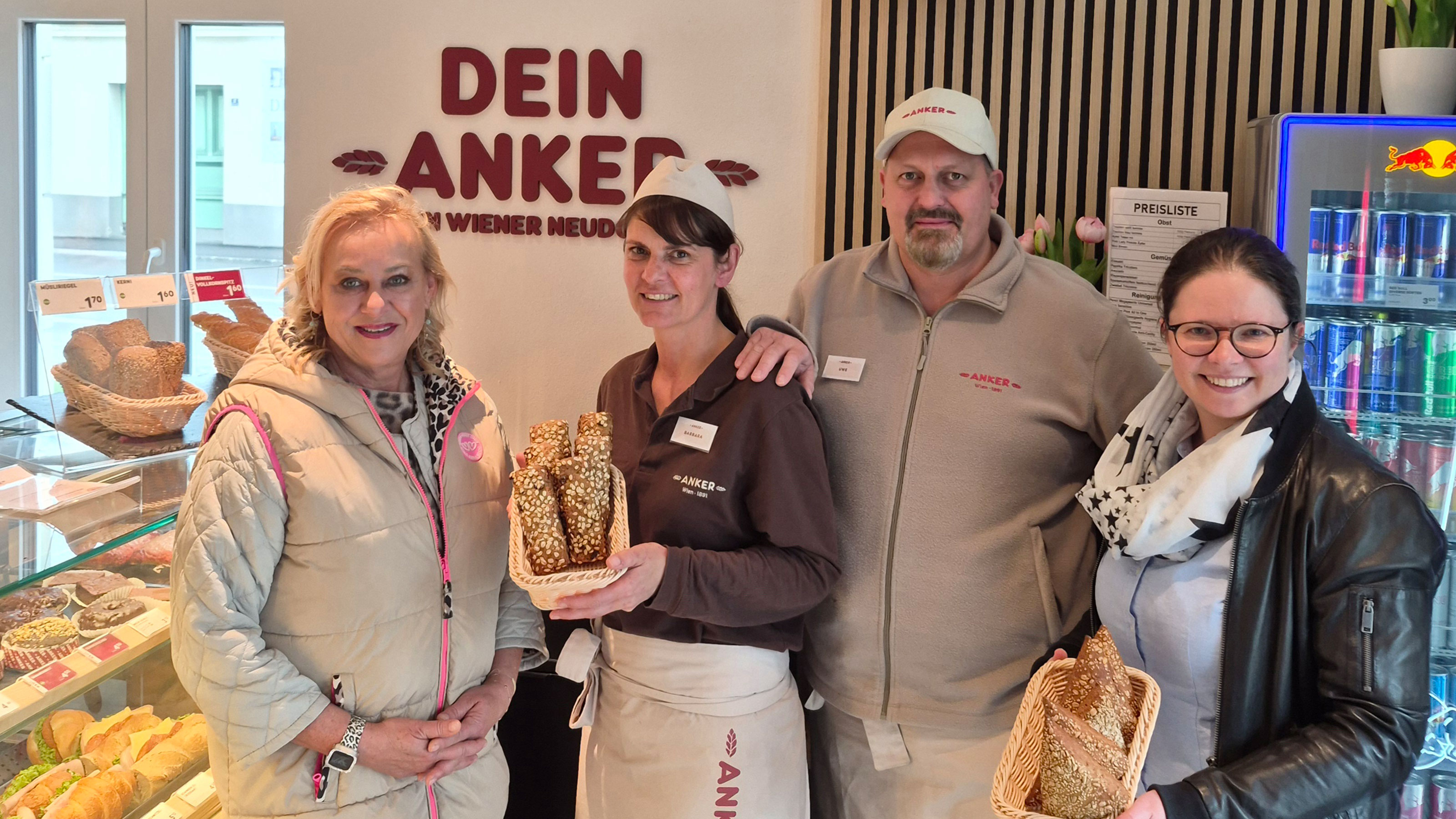 Vier Personen stehen in einer B&auml;ckereifiliale vor einer Verkaufstheke und halten K&ouml;rbe mit l&auml;nglichen, bestreuten Geb&auml;ckst&uuml;cken in den H&auml;nden; im Hintergrund sind Backwaren, ein Getr&auml;nkek&uuml;hlschrank und das Logo &bdquo;DEIN ANKER&ldquo; zu sehen, Gesichter sind unkenntlich gemacht.