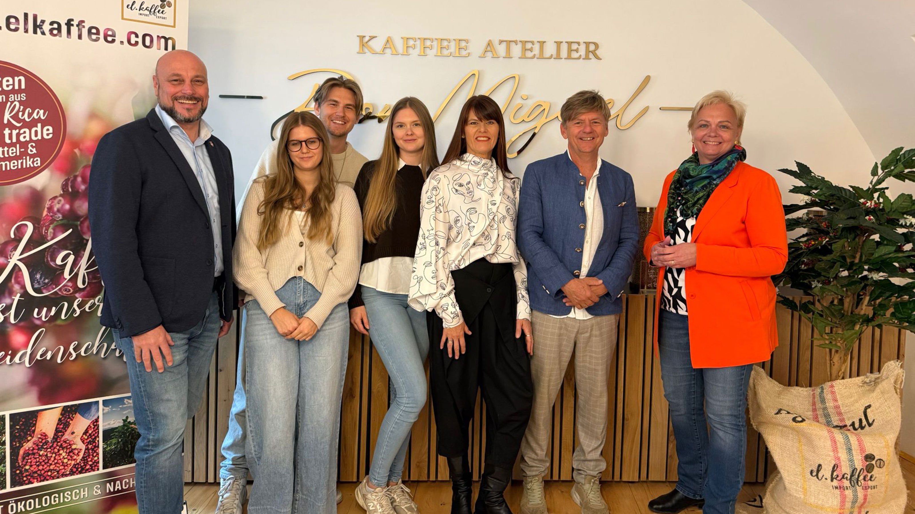 Gruppenfoto von mehreren Personen in einem Kaffee-Atelier vor einer Wand mit der Aufschrift &bdquo;Kaffee Atelier&ldquo;, daneben ein Roll-up mit Kaffeebohnen-Motiv und Markenhinweis.