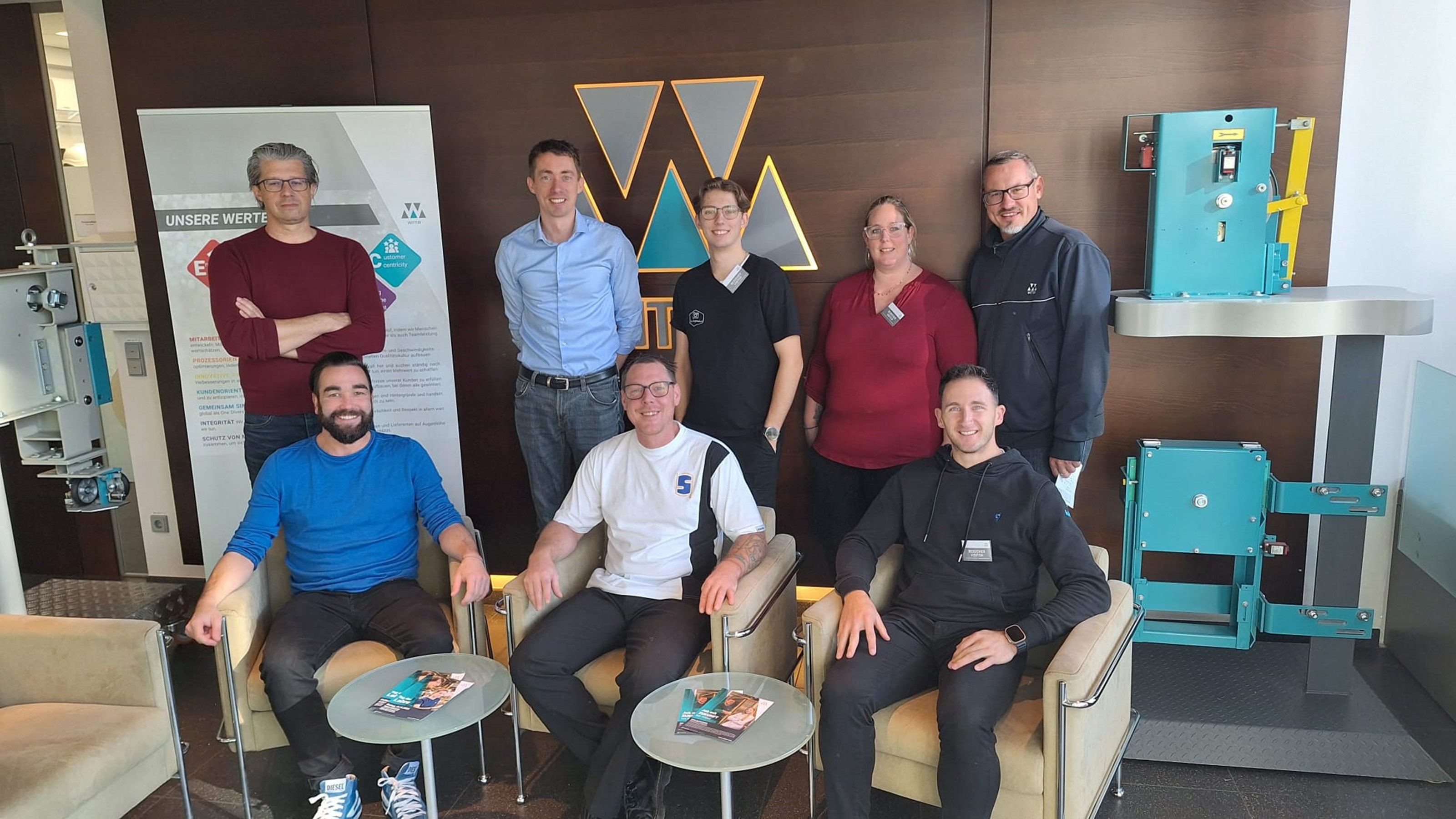 Gruppe von sieben Personen in einem Firmenfoyer, einige sitzen auf Sesseln, andere stehen dahinter. Im Hintergrund sind ein Firmenlogo "Wittur", ein Informationsbanner und Aufzugskomponenten sichtbar.