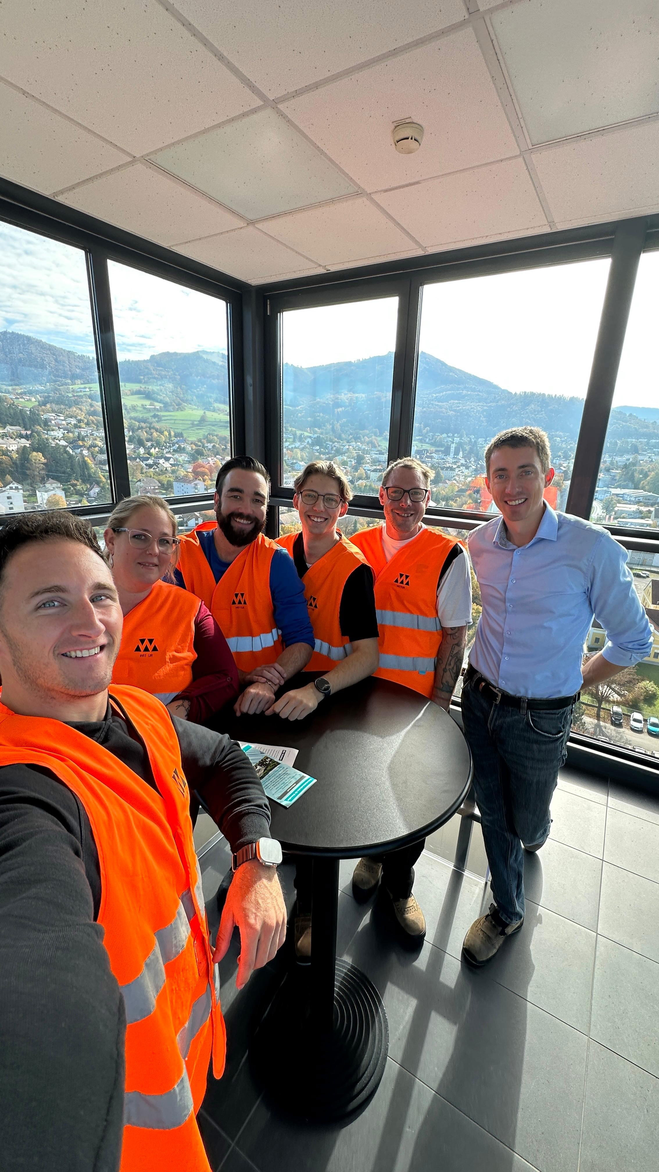 Gruppe von fünf Personen steht in einem verglasten Raum am Turm mit Blick auf eine hügelige Landschaft. Vier tragen orange Warnwesten, eine Person steht daneben in Freizeitkleidung. In der Mitte befindet sich ein runder Stehtisch mit Informationsmaterial.