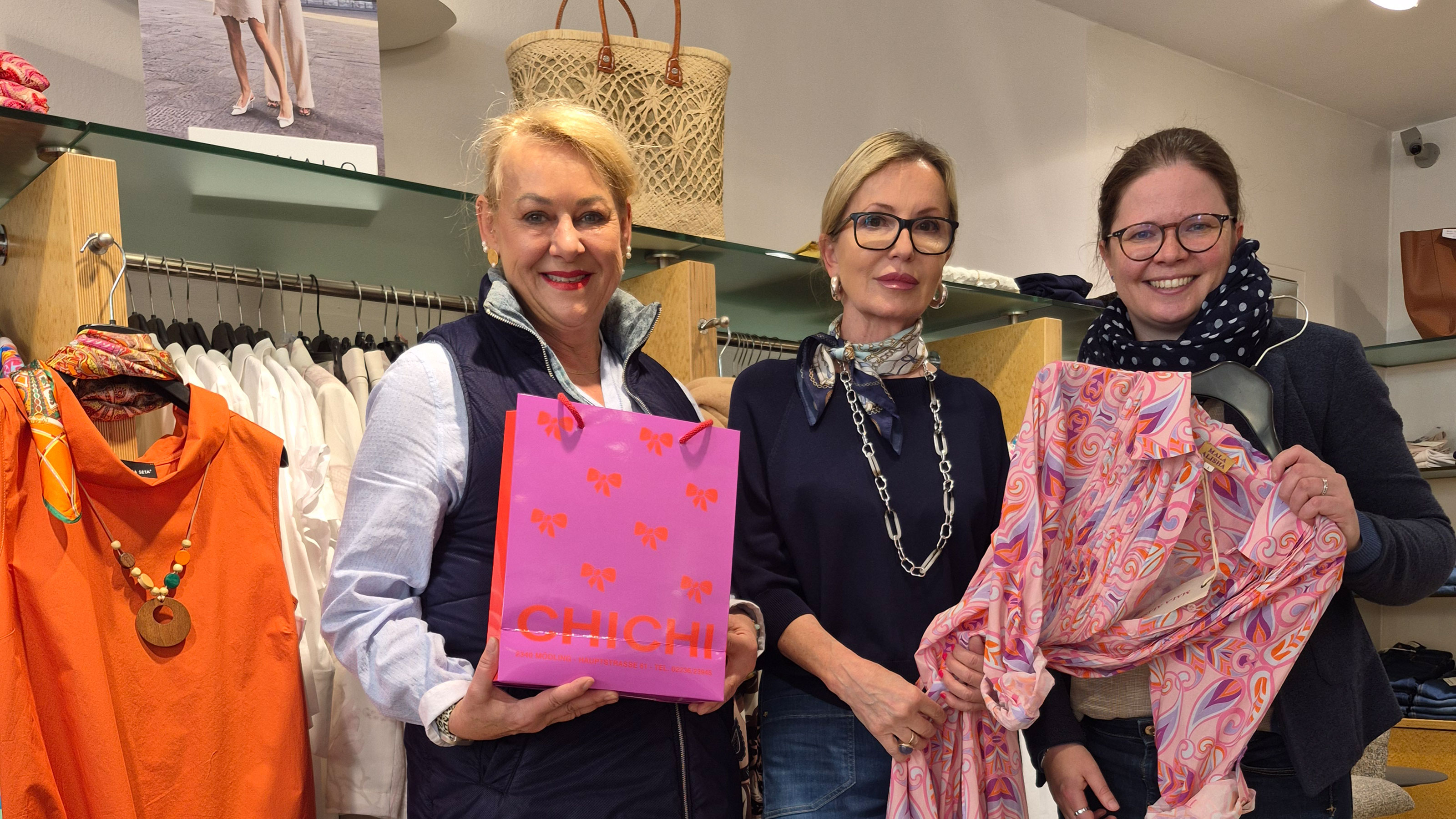 Drei Personen stehen in einer Boutique zwischen Kleiderst&auml;ndern und pr&auml;sentieren eine Einkaufstasche sowie ein Kleidungsst&uuml;ck.