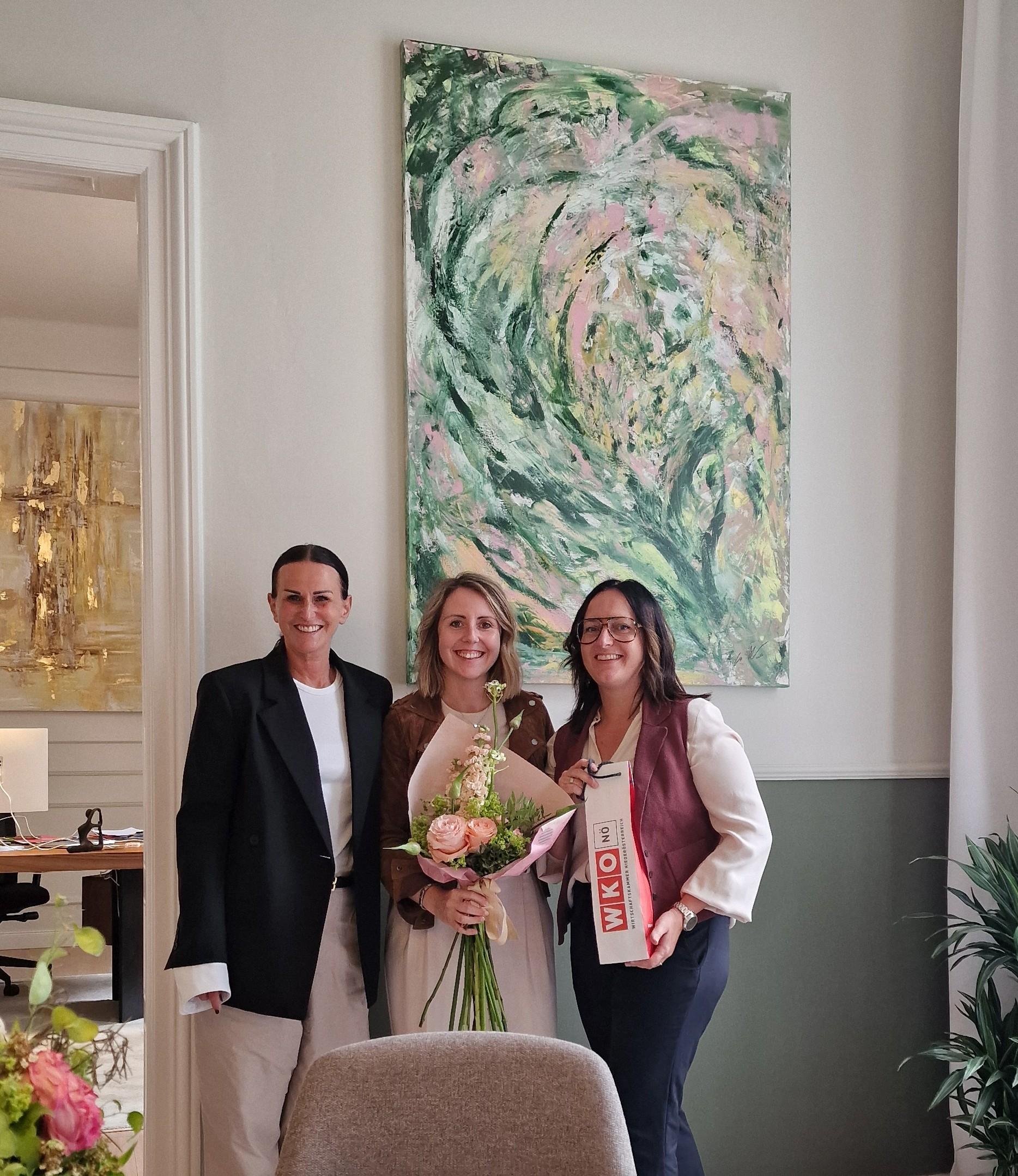 Drei Frauen stehen l&auml;chelnd in einem hellen Raum vor einem gro&szlig;en abstrakten Gem&auml;lde. Die Frau in der Mitte h&auml;lt einen Blumenstrau&szlig;, die Frau rechts ein Geschenk.