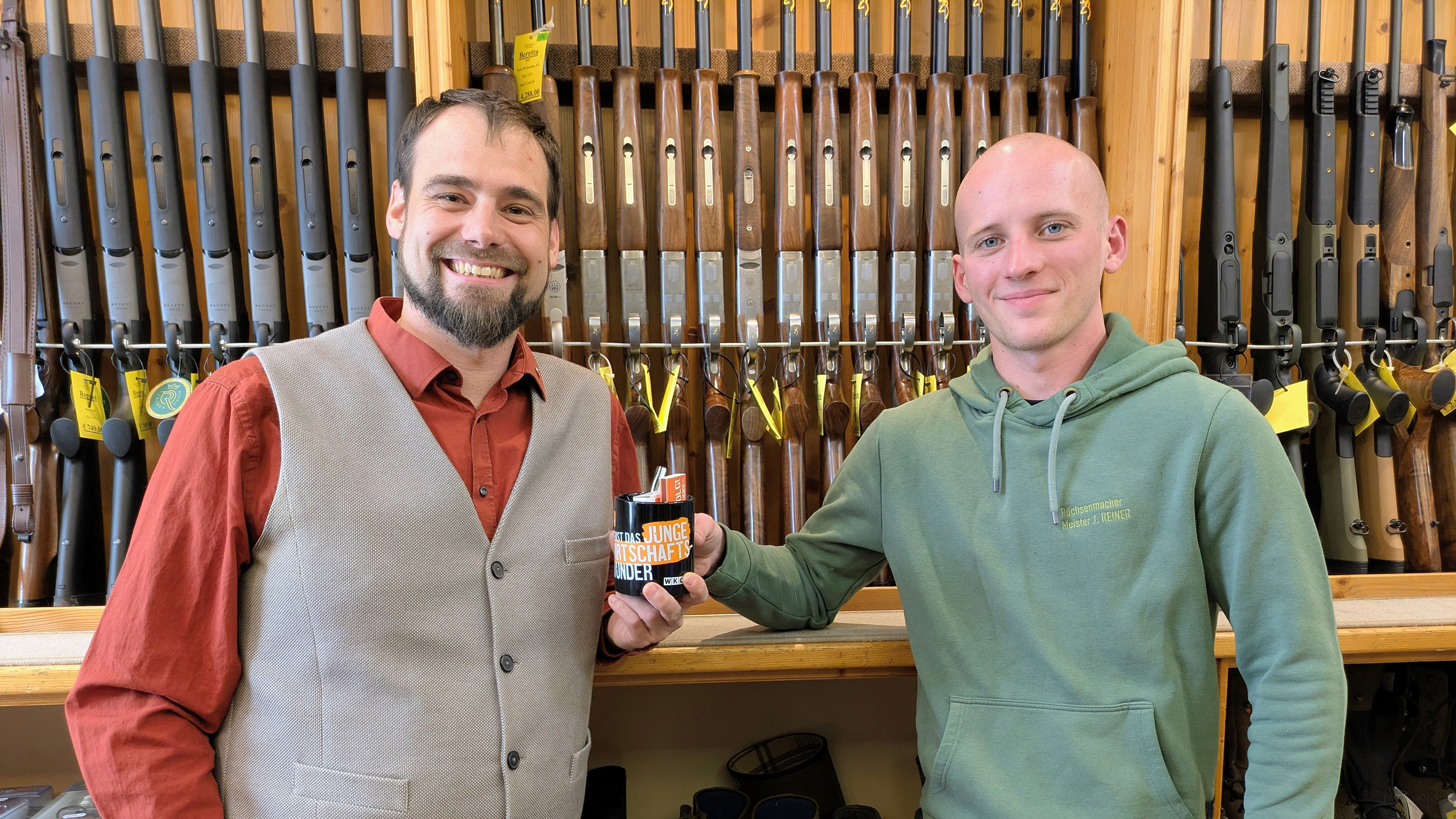 Zwei Personen stehen in einem Geschäft vor einer Wand mit Jagd- und Sportgewehren und halten gemeinsam eine schwarzes Häferl mit Aufdruck "Junge Wirtschaft" in der Hand.
