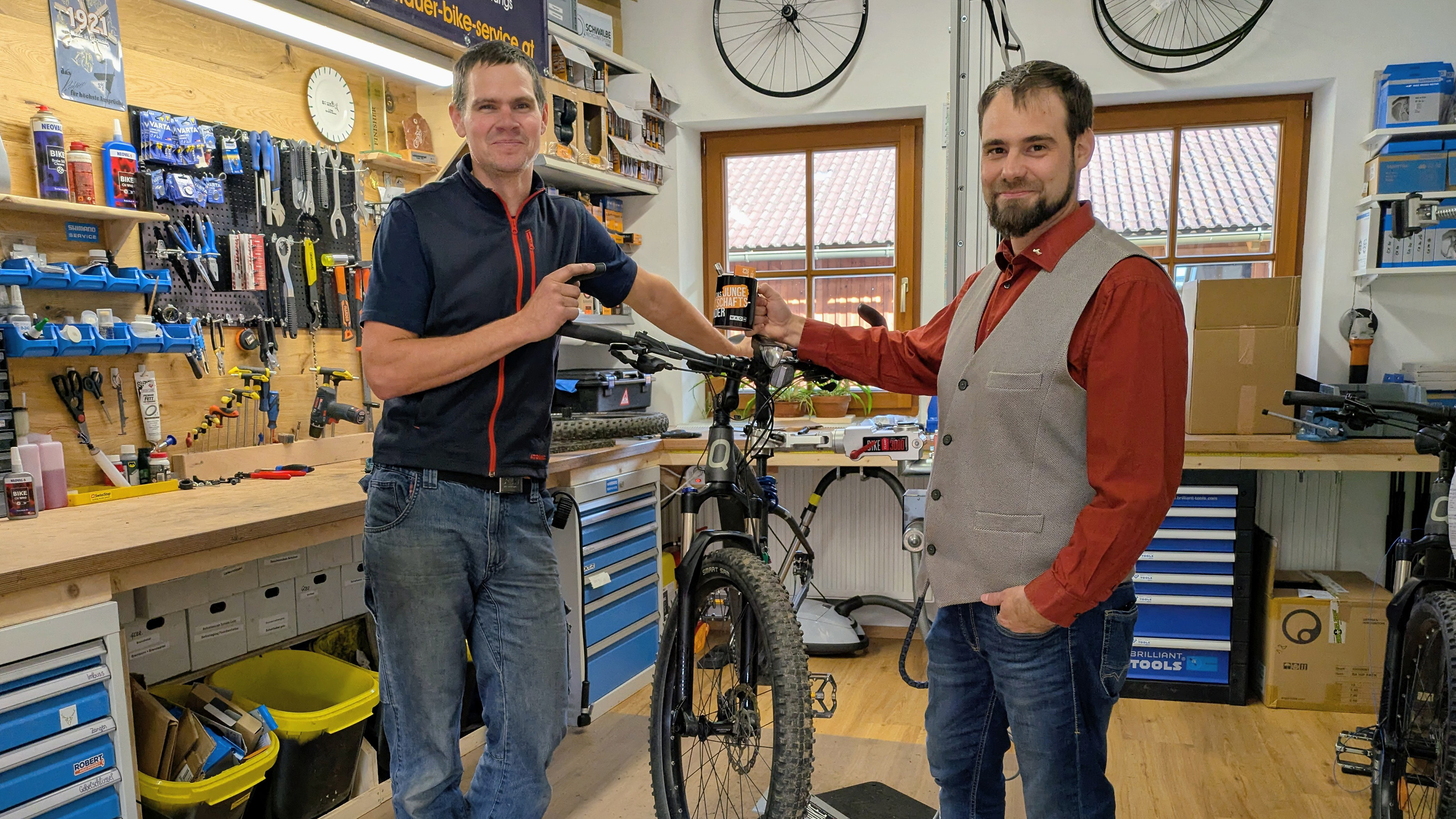 Zwei Personen stehen in einer Fahrradwerkstatt neben einem Mountainbike. Im Hintergrund sind Werkbänke mit Werkzeugen, Ersatzteilen und Regalen sichtbar.