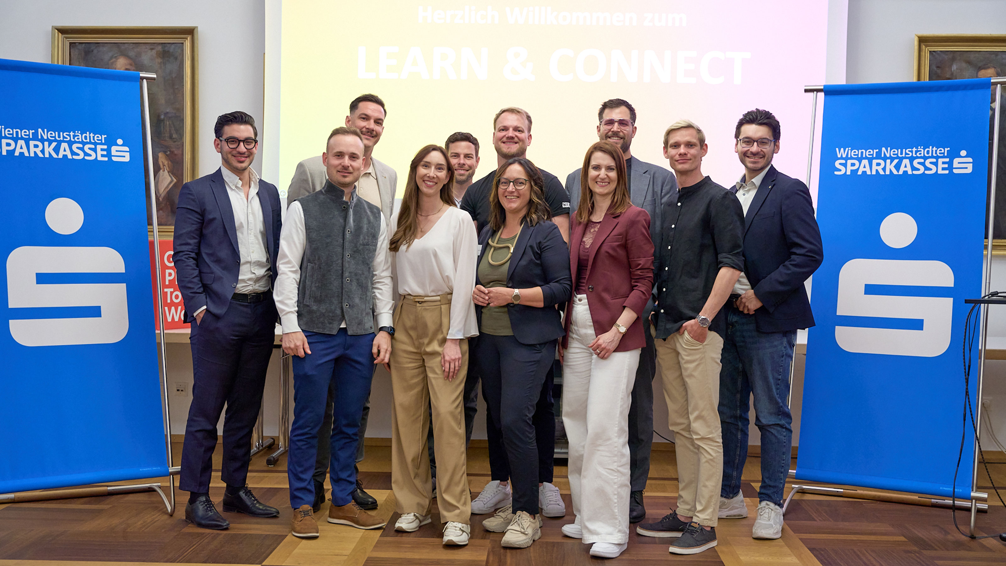 Gruppenfoto von mehreren Erwachsenen bei einer Indoor-Veranstaltung; die Gruppe steht zwischen zwei blauen Roll-ups der Wiener Neust&auml;dter Sparkasse, im Hintergrund eine Pr&auml;sentationsfolie mit der Aufschrift &bdquo;Herzlich Willkommen zum LEARN & CONNECT&ldquo;.