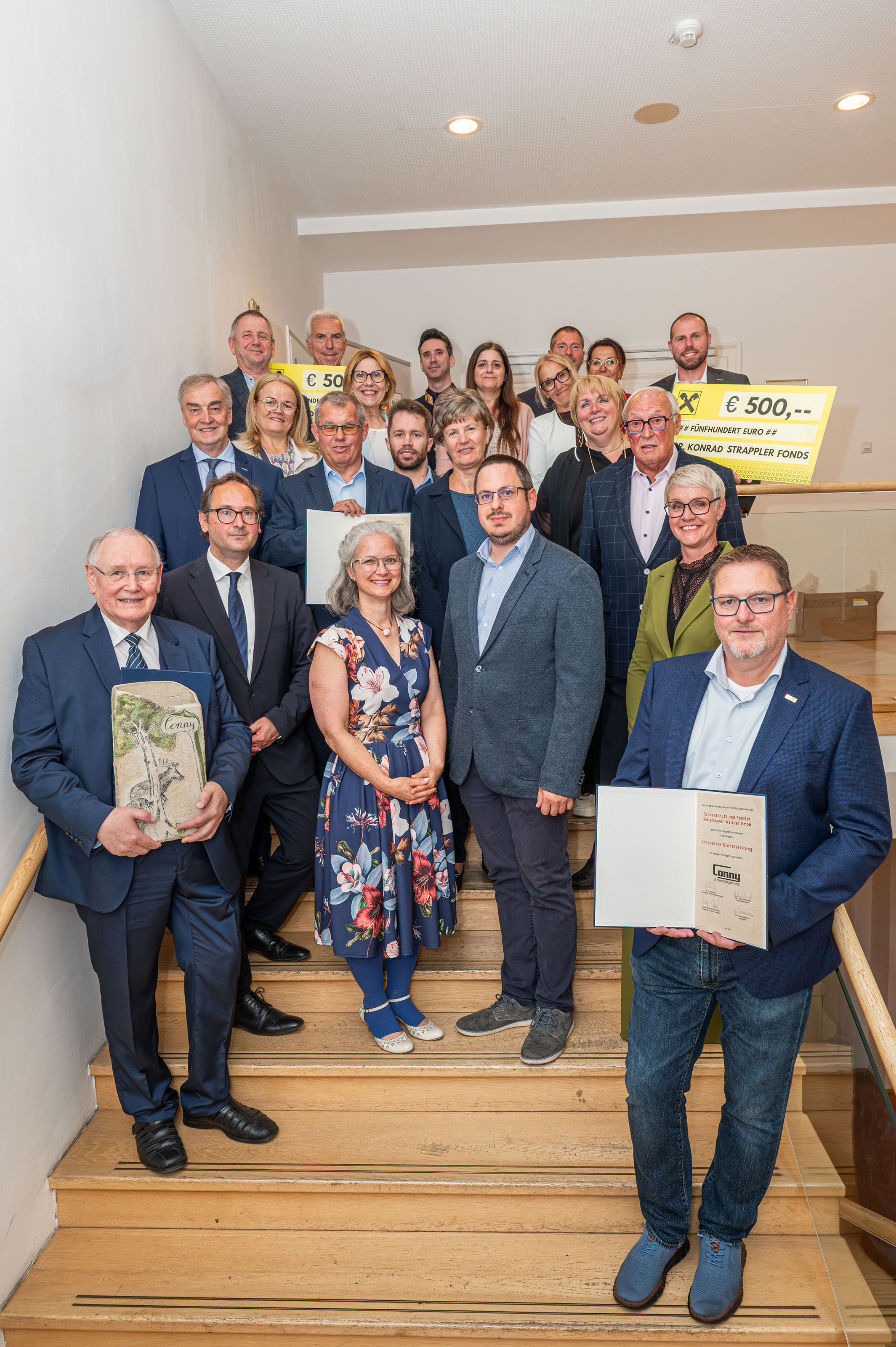 Gruppenfoto von rund 18 Personen auf einer Treppe in einem Innenraum; mehrere Personen halten Urkunden sowie zwei gro&szlig;e Tafeln mit der Aufschrift &bdquo;&euro; 500,--&ldquo;.
