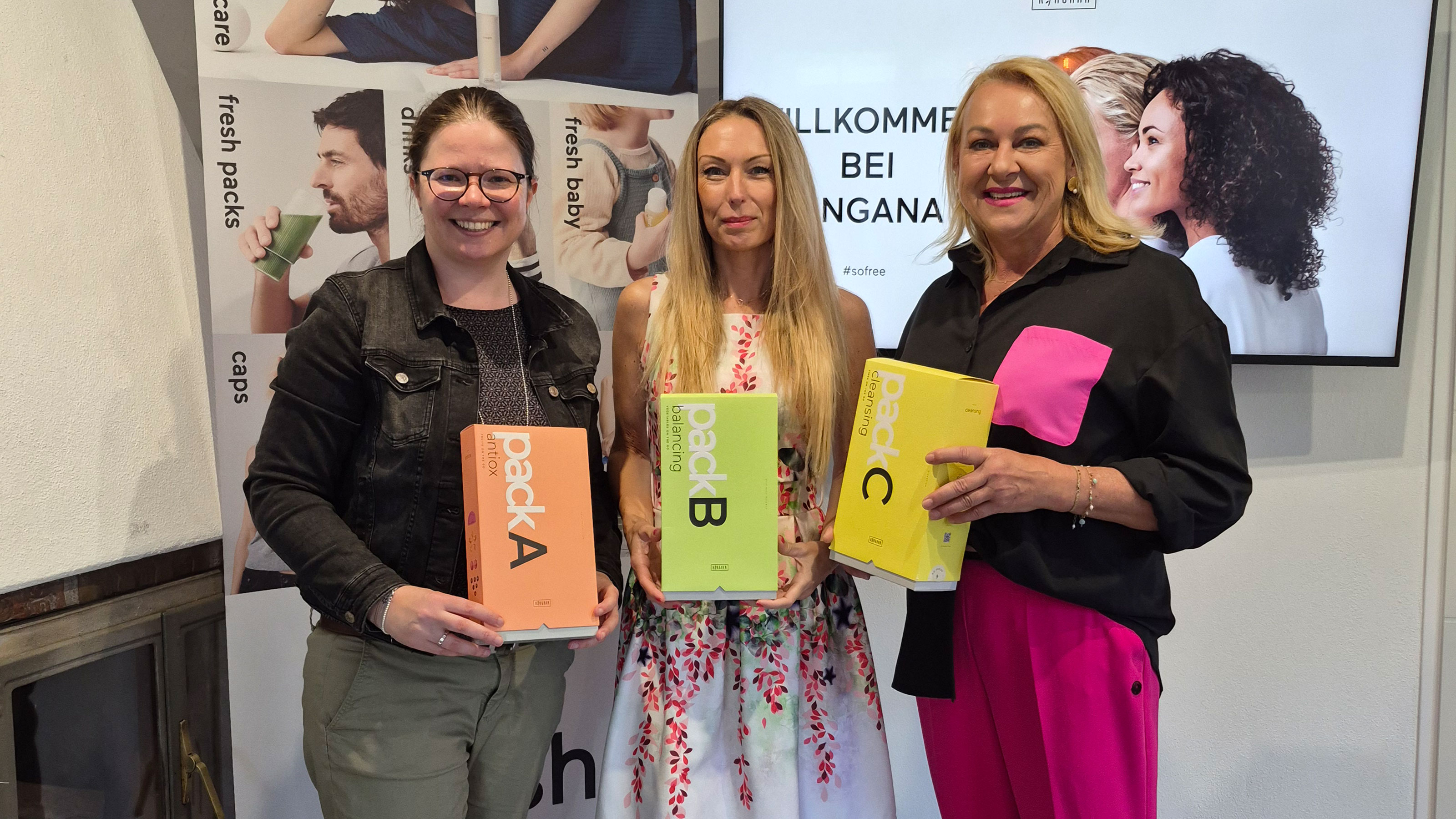 Drei Personen stehen in einem Innenraum vor einem Ringana‑Roll-up und einem Bildschirm mit der Aufschrift &bdquo;Willkommen bei Ringana&ldquo; und halten farbige Produktboxen mit den Bezeichnungen Pack A, Pack B und Pack C in den H&auml;nden.