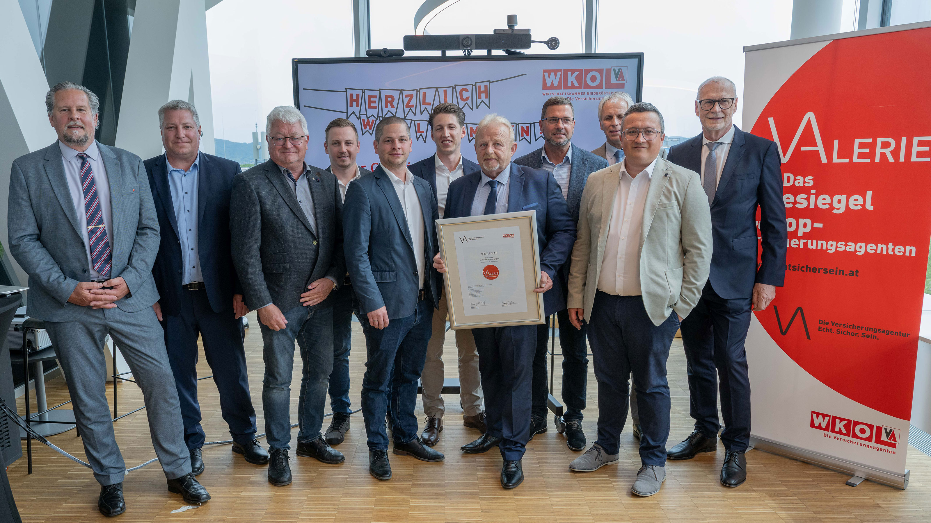 Gruppenfoto von mehreren Personen in Businesskleidung bei einer Auszeichnungsveranstaltung, eine Person h&auml;lt eine gerahmte Urkunde; im Hintergrund sind WK&Ouml;‑Logos und ein rotes &bdquo;VALERIE &ndash; Das G&uuml;tesiegel f&uuml;r Top‑Werbungsagenten&ldquo;-Banner zu sehen.