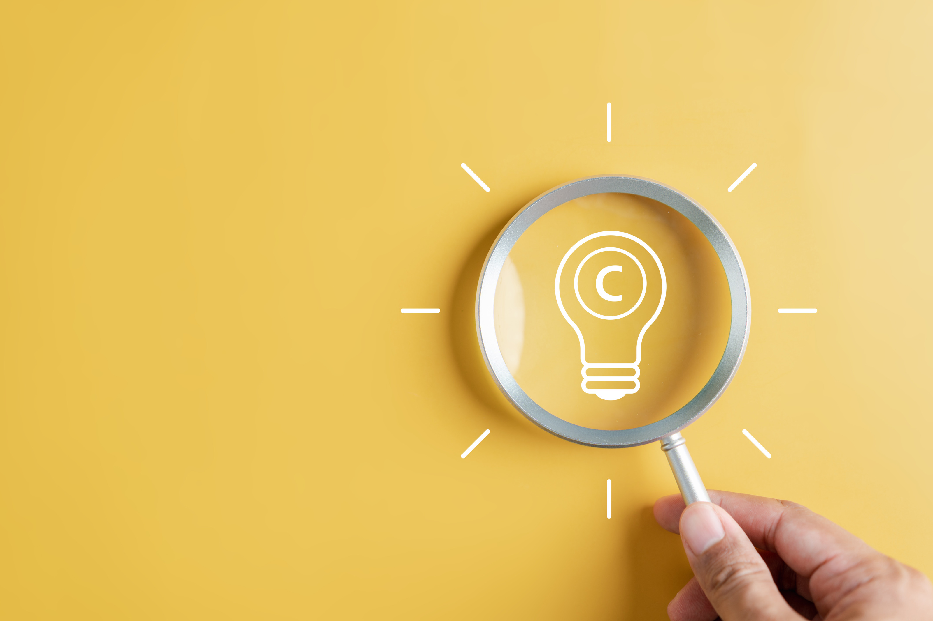 Nahaufnahme einer Lupe, die von der Hand einer Person gehalten wird. Die Lupe fokussiert das Symbol einer Glühbirne, um die  Lupe herum sind vertikale Striche. In der Glühbirne ist ein C in einem Kreis als Zeichen für Copyright. 