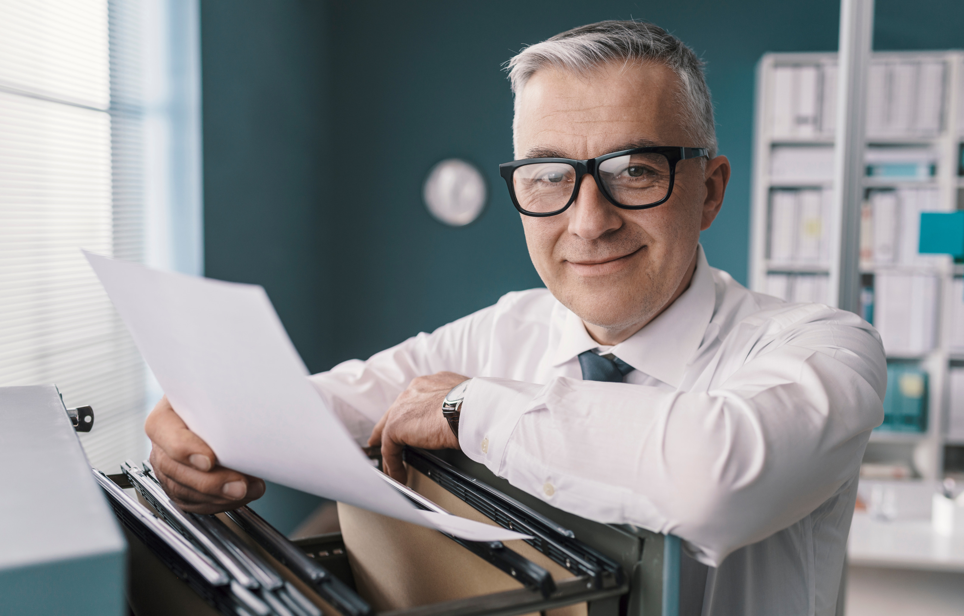 Sanft lächelnde Person mit Brille lehnt über Aktenlade und hält Dokument in einer Hand