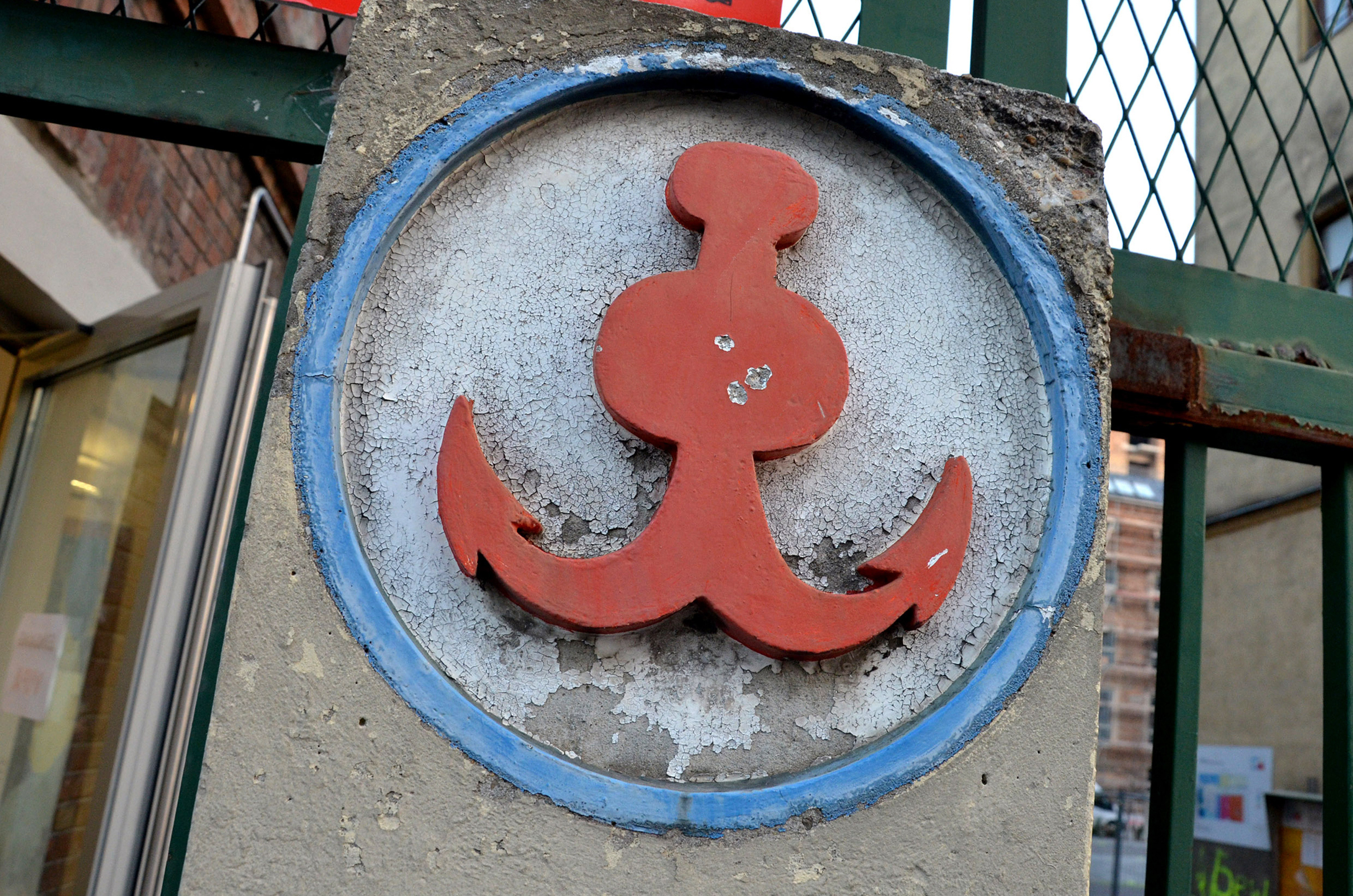 Wandrelief auf Betonstrebe eines Zauns: Symbol eines roten Ankers mit blauer, kreisförmiger Umrandung