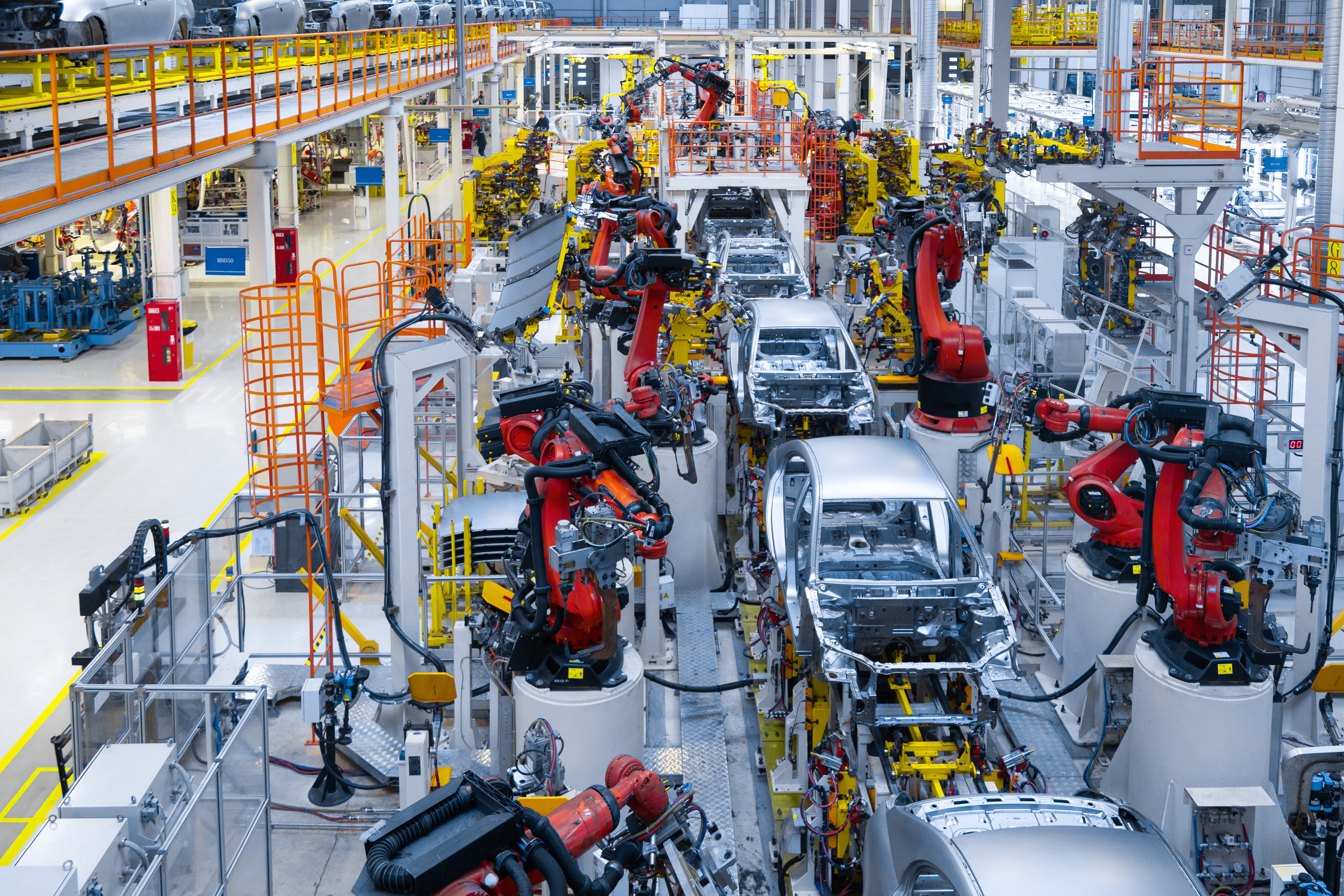 Top-Shot einer gro&szlig;en Fabrik, in der die H&uuml;lle von grauen Autos sind. Links und rechts der Autos sind gro&szlig;e, rote Roboterarme, die auf grauen, hohen Plattformen stehen.