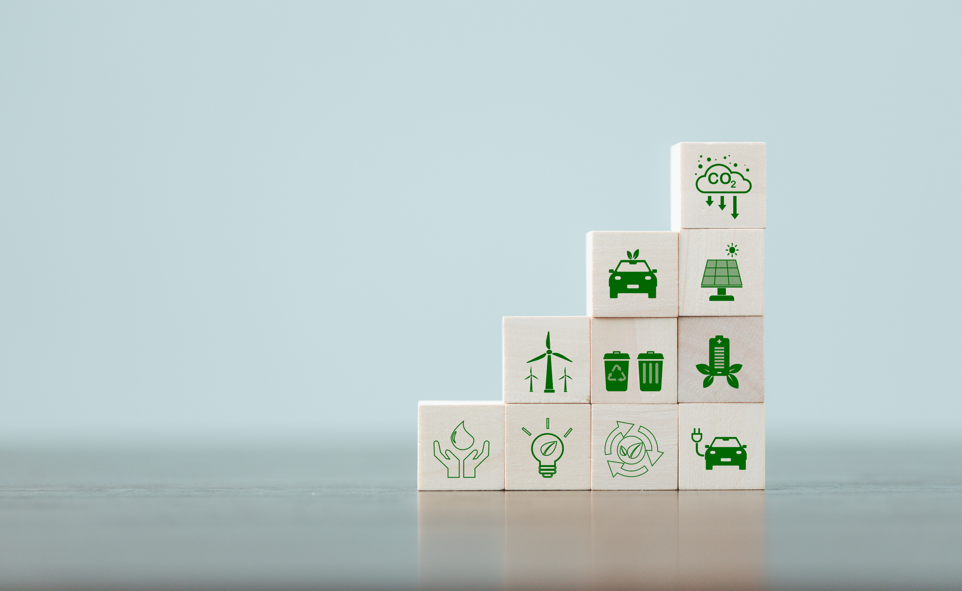 Mehrere Holzw&uuml;rfel aufeinander gestapelt auf einer hellen Oberfl&auml;che mit gr&uuml;nen Symbolen zum Thema Klimaschutz: Eine Wolke mit der Aufschrift CO2, Autos, Solar-Panel, Windr&auml;der, Mistk&uuml;bel, Gl&uuml;hbirne, Pfeile, Handfl&auml;chen mit Wassertropfen.