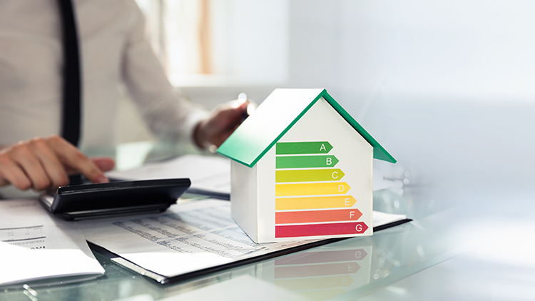 Minitaturhaus mit Abbildung der Energieklassen, im Hintergrund Person auf Taschenrechner tippend