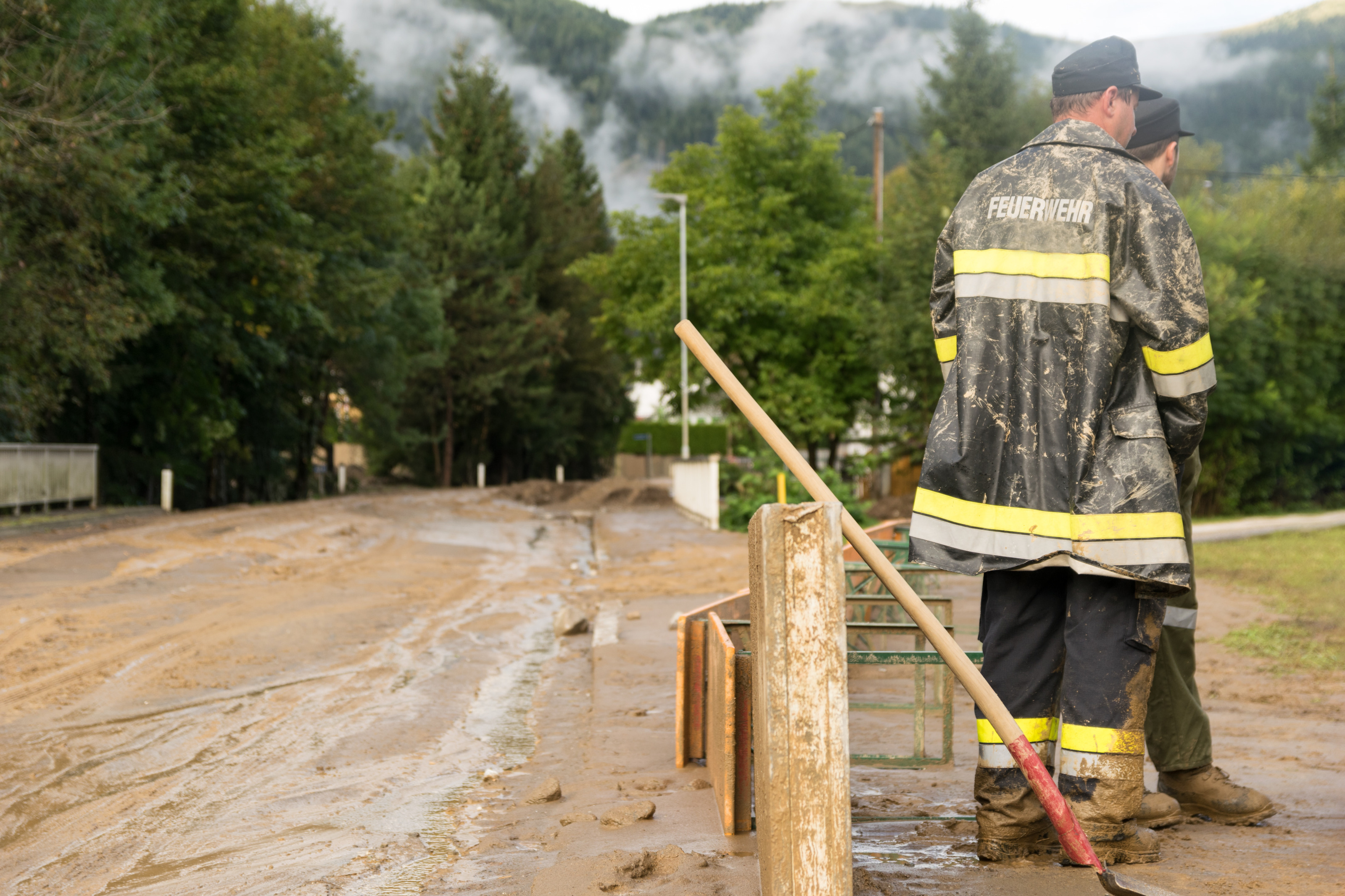 Zwei Personen in R&uuml;ckenansicht in Schutzkleidung mit der Aufschrift Feuerwehr stehen neben wasserverschlammter Stra&szlig;e, die von B&auml;umen ums&auml;umt ist, neben den Personen lehnt eine Schaufel an Holzbalken.
