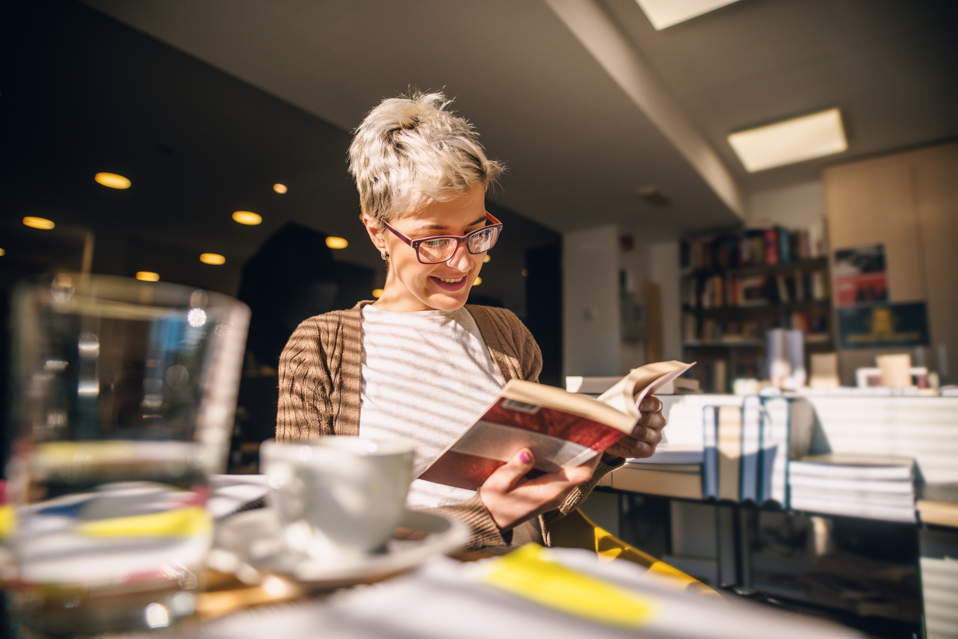 L&auml;chelnde Person mit Brillen sitzt an Tisch und h&auml;lt Buch in H&auml;nden auf das sie blickt, im Hintergrund verschwommen Tische mit B&uuml;cherstapeln und volle B&uuml;cherregale